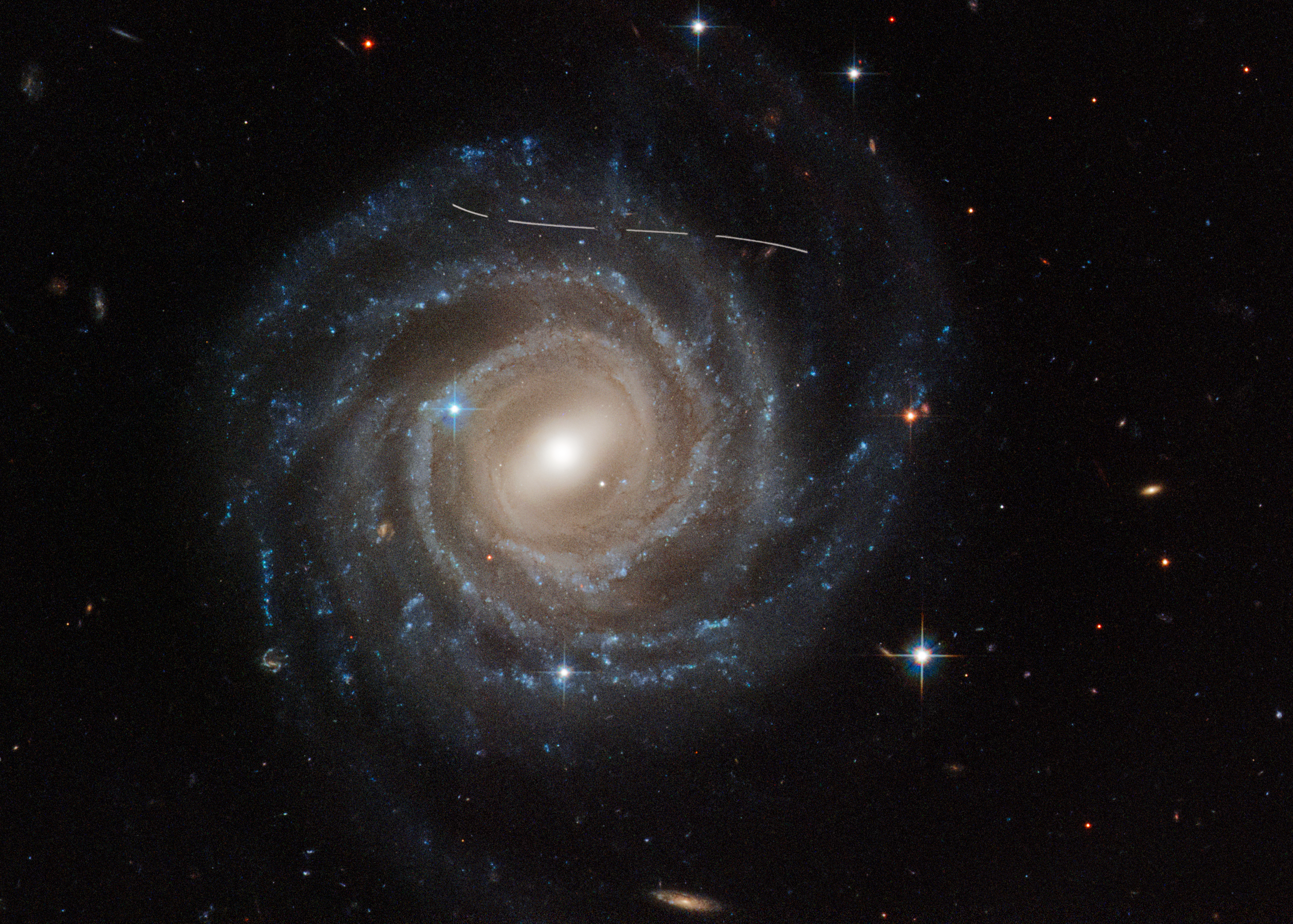 This is a Hubble Space Telescope image of the barred spiral galaxy UGC 12158. The majestic galaxy has a pinwheel shape made up of bright blue stars wound around a yellow-white hub of central stars. The hub has a slash of stars across it, called a bar. The galaxy is tilted face-on to our view from Earth. A slightly s-shaped white line across the top is a Hubble image is of an asteroid streaking across Hubble's view. It looks dashed because the image is a combination of several exposures of the asteroid flying by like a race car.