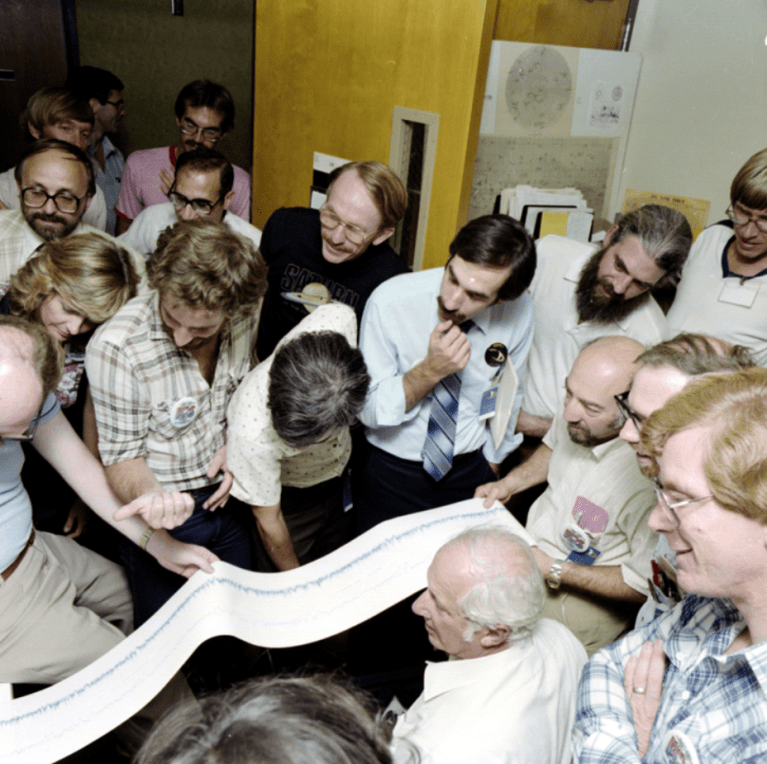 A group of scientists looking down at a long ticker tape of data