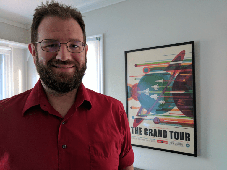 A man standing proudly next to a poster on the wall that reads "The Grand Tour"