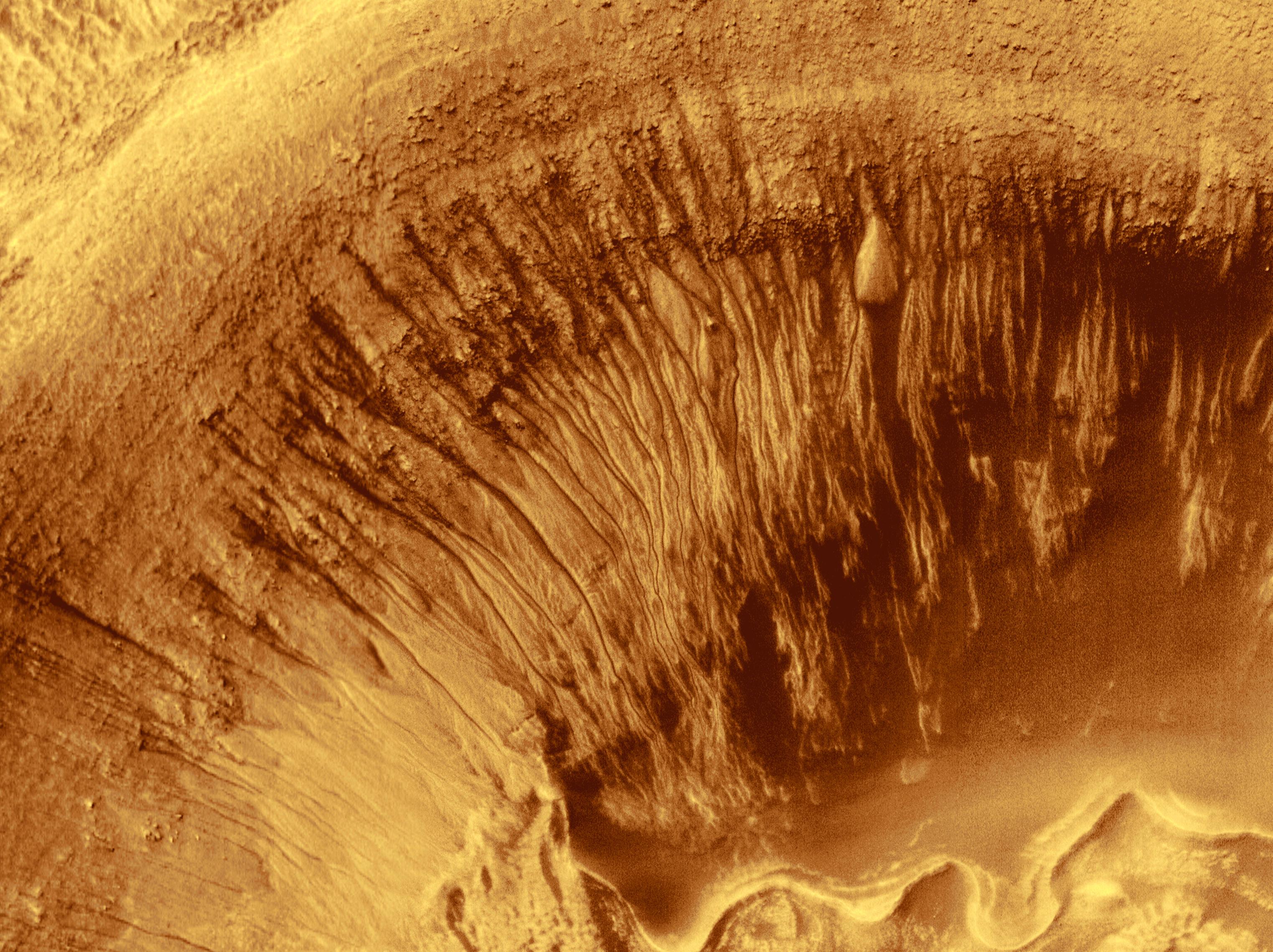 This overhead view of a Martian crater shows the upper-left quarter of the crater – its rim and the walls descending to the floor below. The rim and floor appear as a bright yellow-orange, while the wall is in various darker shades culminating in a deep rust color at the bottom where it meets the wall. The rim is fairly smooth while the walls are grooved with straight and slightly curved lines until they meet the crater floor, making the scene look like the underside of a mushroom. The crater floor is somewhat uneven, highlighted by a ribbon of bright yellowish-orange terrain, undulating from the middle of the bottom frame of the image toward the right edge of the frame, where it ends at in the lower quarter.