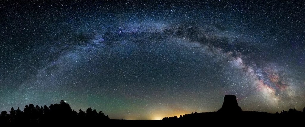 The disk of the Milky Way makes a blue bowl-shaped arc over the land and trees.