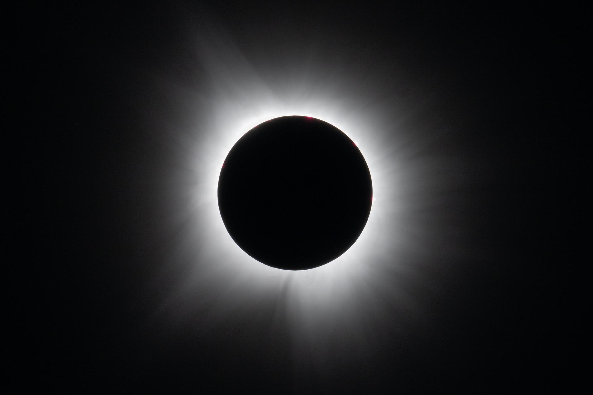 Against a black background, the total solar eclipse. It is a black circle surrounded by white, wispy streams of light that flow away from the black circle in every direction.