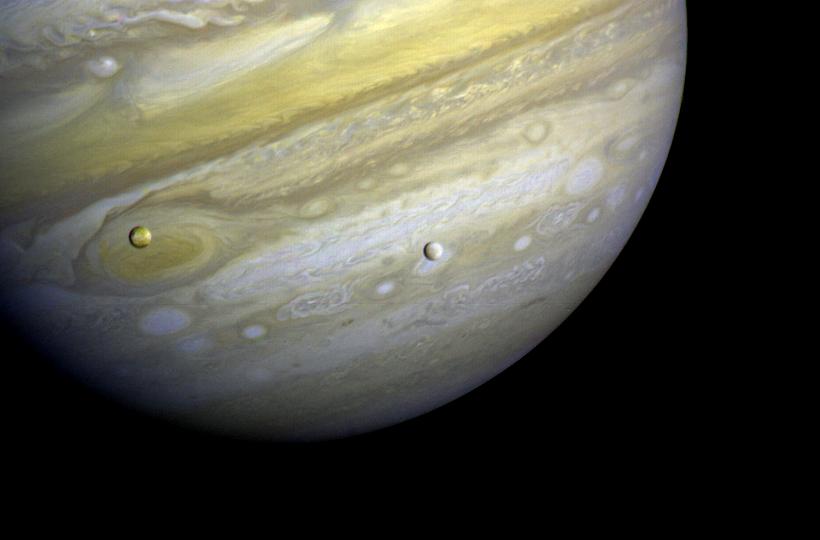 Two moons cross the face of Jupiter, which is ringed with bands of clouds and storms.