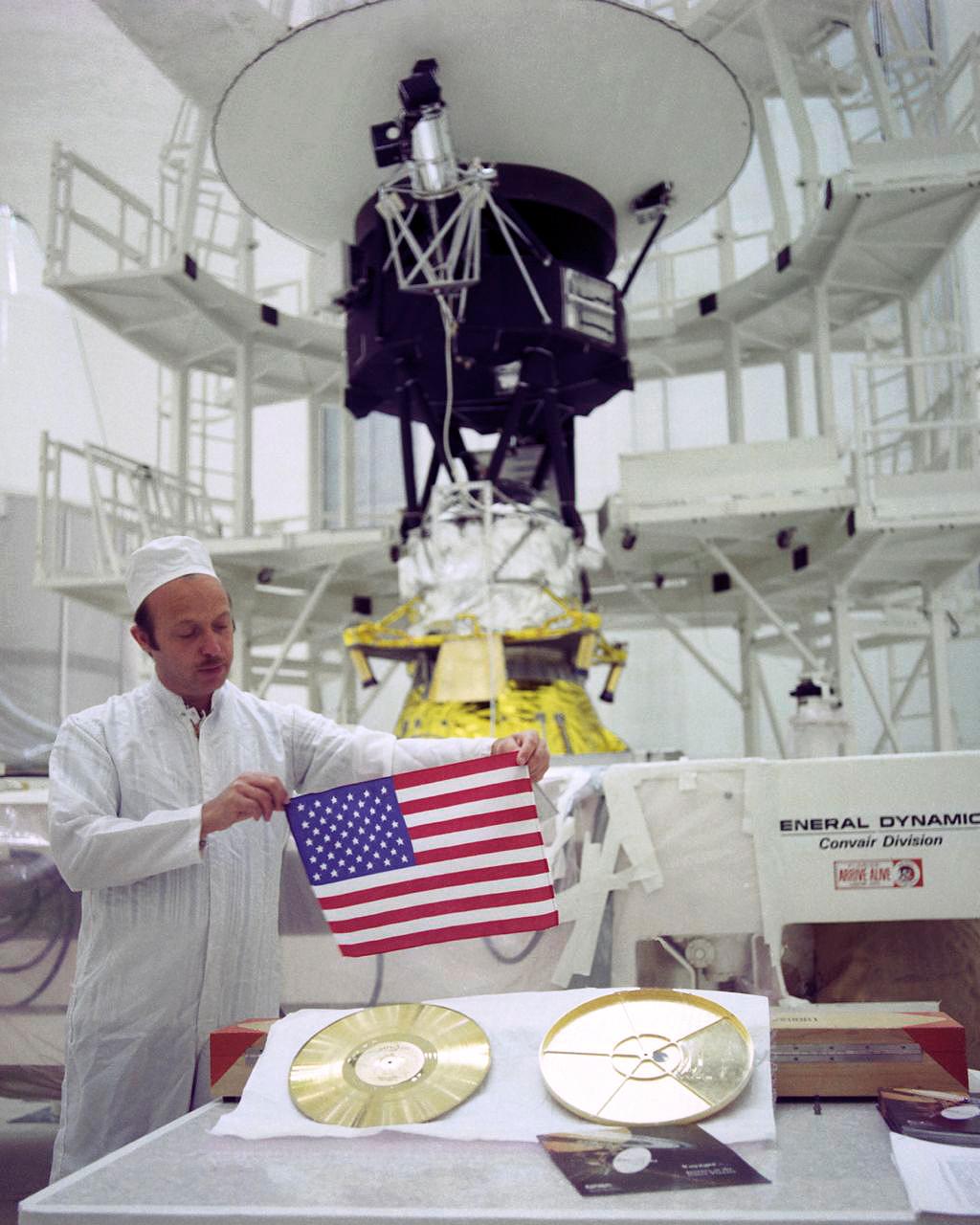 John Casani, Voyager project manager in 1977, holding a small Dacron flag that was folded and sewed into the thermal blankets of the Voyager spacecraft before they launched 36 years ago. Below him lie the Golden Record (left) and its cover (right). In the background stands Voyager 2 before it headed to the launch pad.