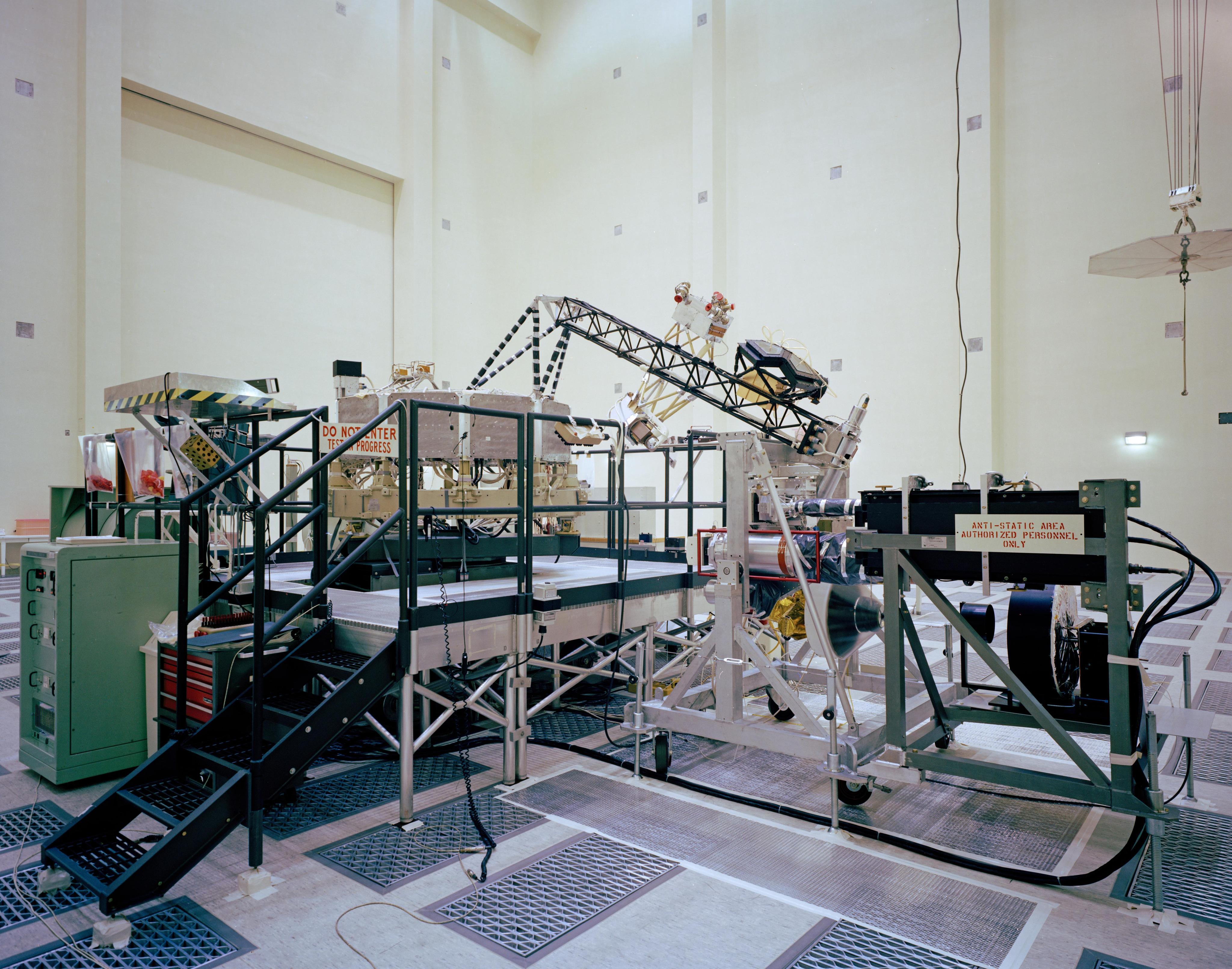 System test configuration for Voyager. The spacecraft's 10-sided bus is visible behind the catwalk railing in the foreground. The boom that holds several of the spacecraft's science instruments arches above the railing.
