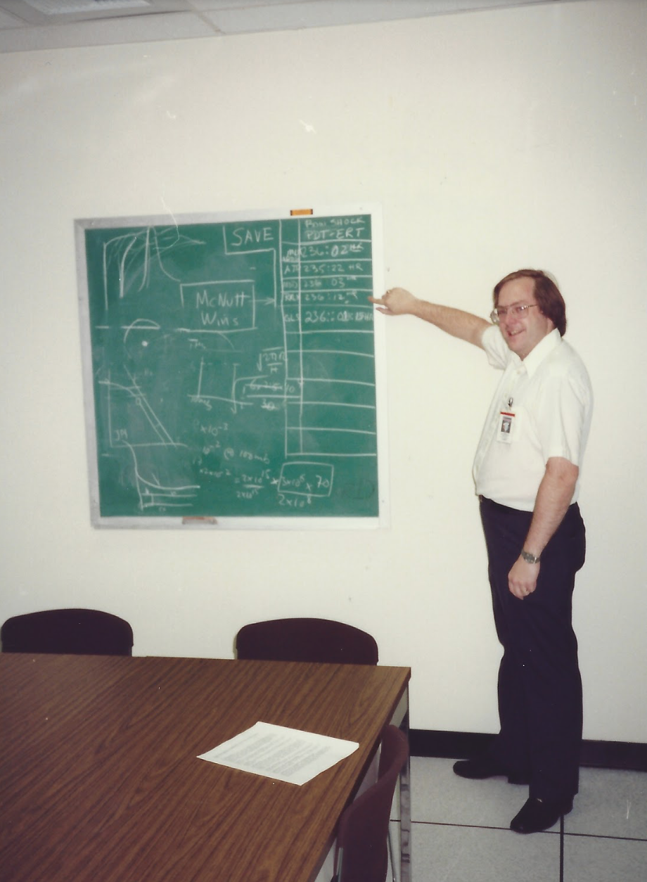 A man standing next to a chalkboard, pointing at figures written on it