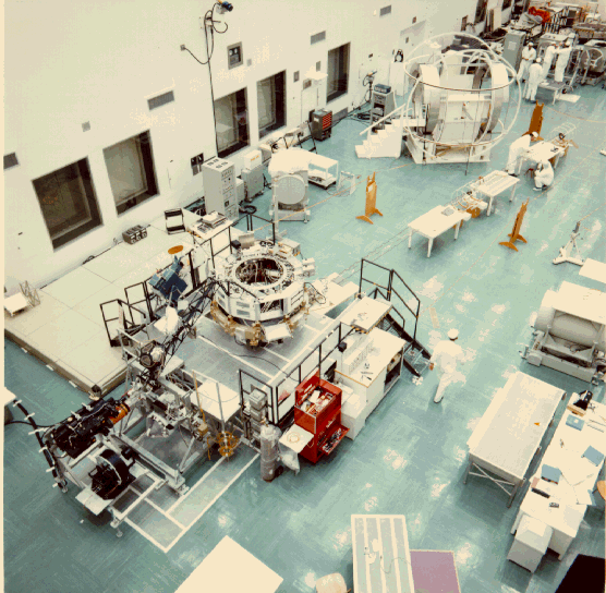 A high view shows parts of a spacecraft being assembled on a clean room floor.