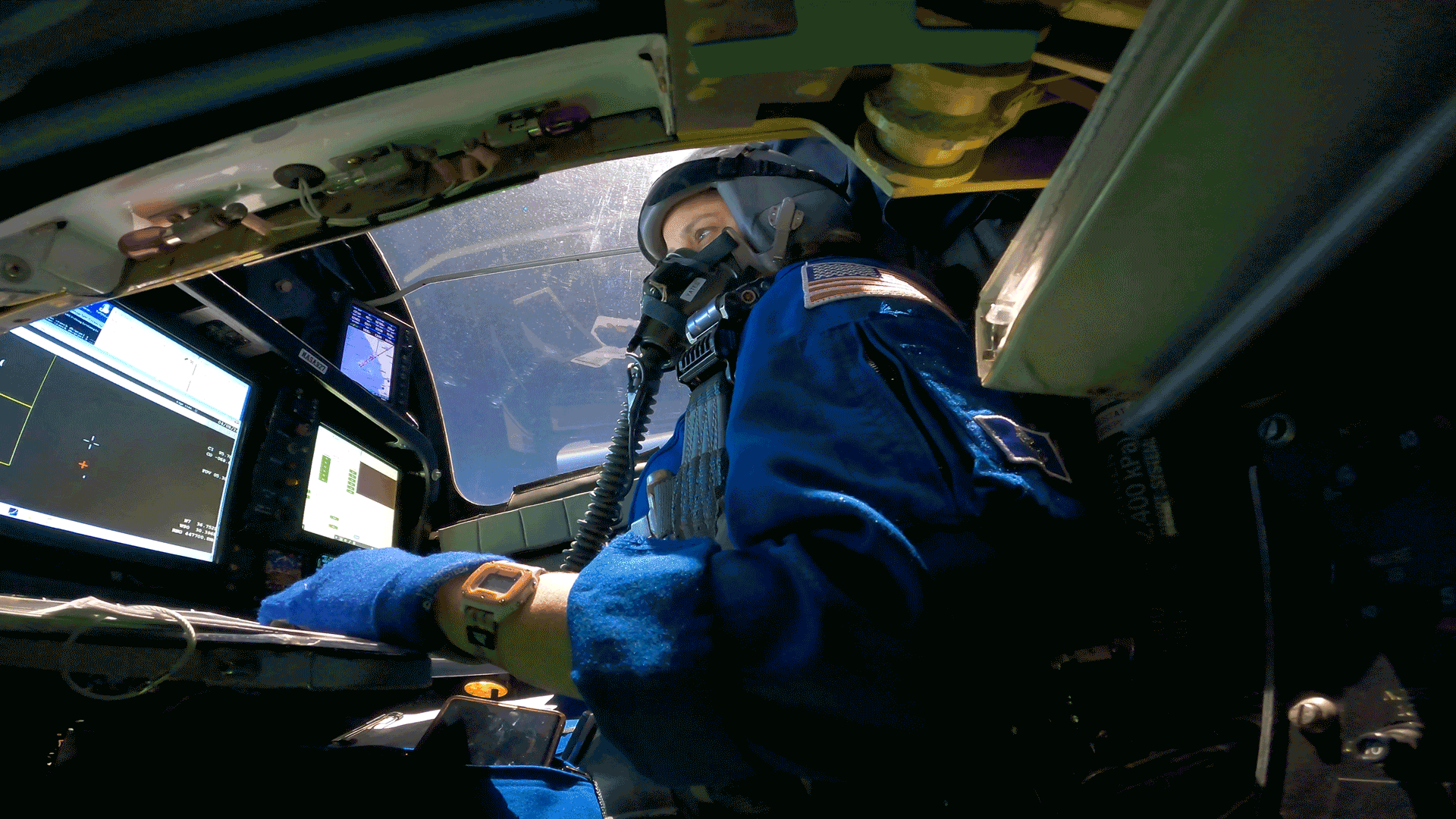 While flying, a view of the pilot in the cockpit from below her seat. The pilot is wearing a blue suit and a helmet with a tube attached to the front. In front of her are several screens, including one showing the total solar eclipse. The cockpit gets darker and darker as she flies in the eclipse's shadow.