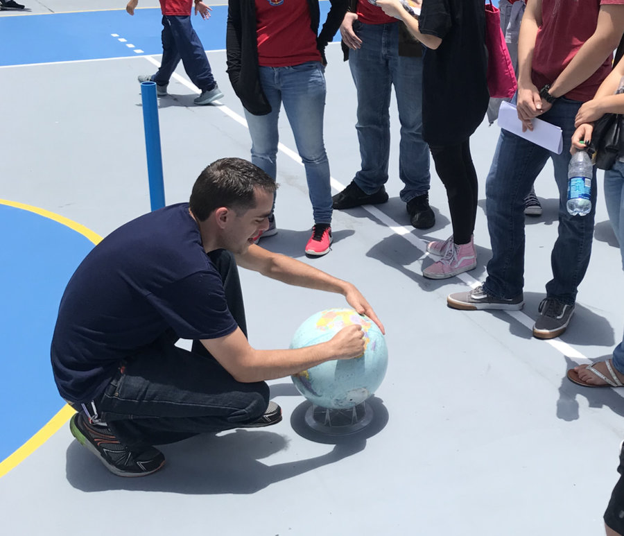 A man squats on the ground surrounded by people, and only their legs are visible. He is pointing to a spot on a globe of the Earth which is propped above the ground on a small plastic stand. Shadows below everything in the picture are very small.