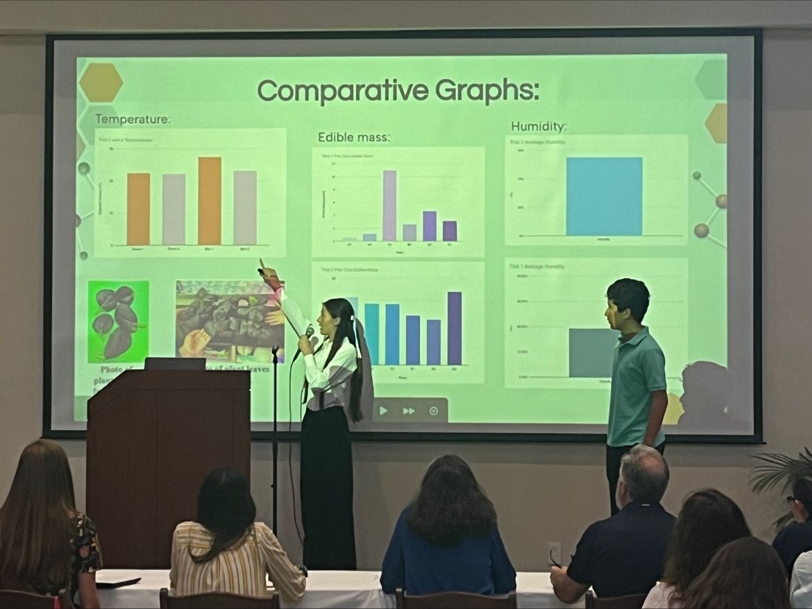A student speaks into a microphone while pointing up at the projector screen behind them showing comparative graphs of their research results. Another student presenter stands near them on the stage.