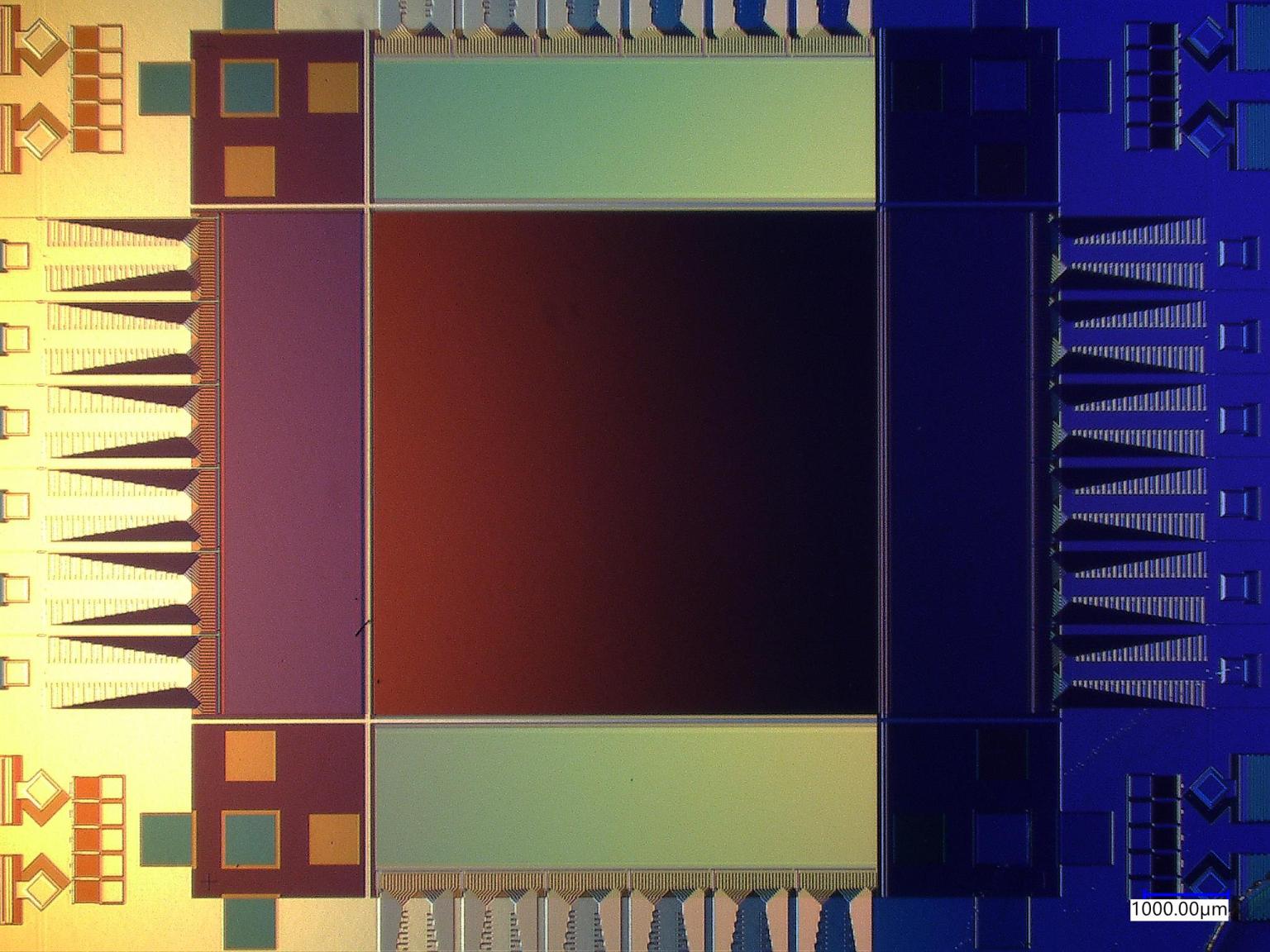 A microscope image of a rectangular chip, showing the different parts of the superconducting camera, including imaging area and ancillary electronics. The chip has a colorful patina, with hues of yellow on the left, red in the center, and blue on the right.