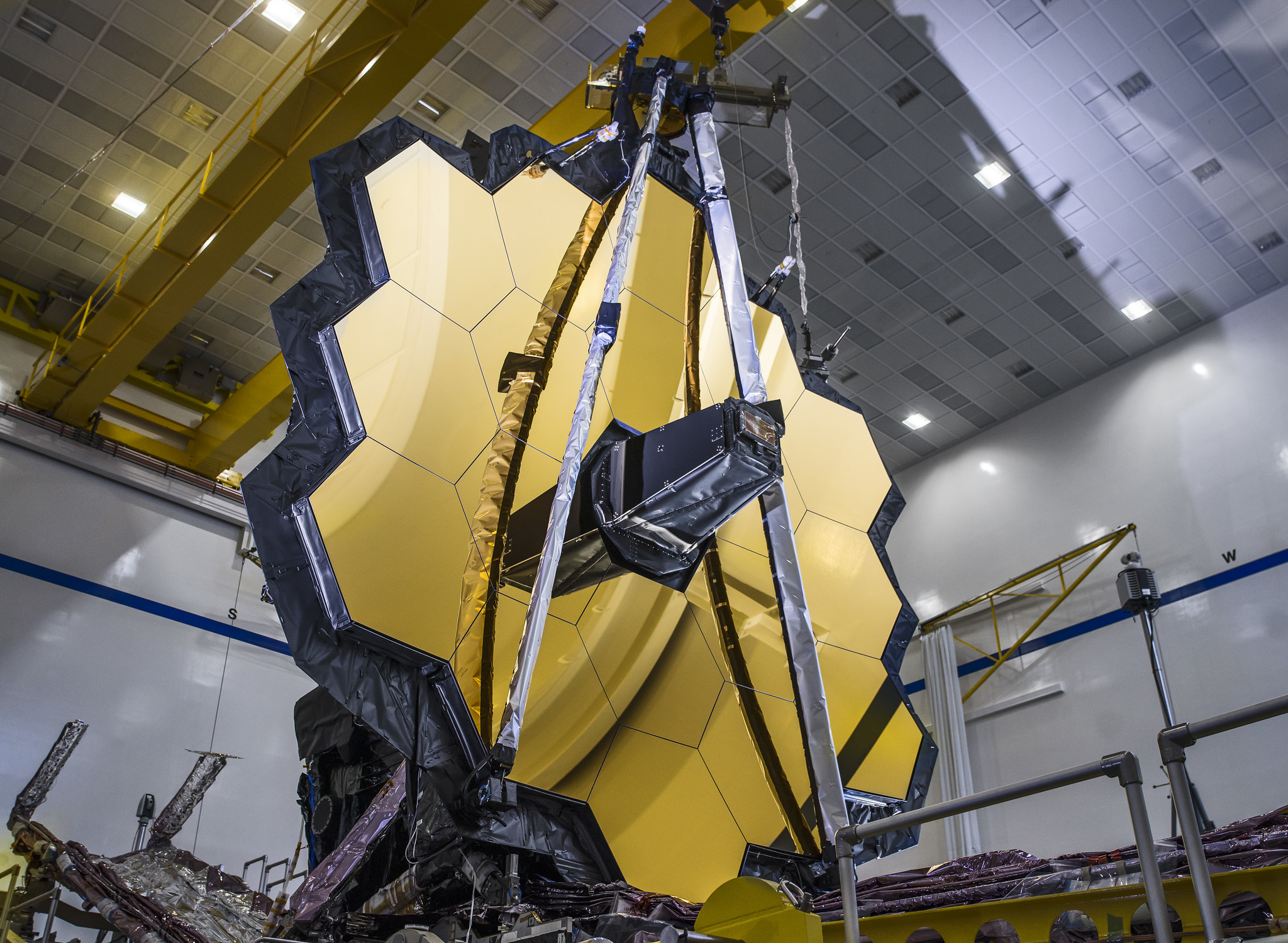 Webb’s giant golden mirror stands tall. Light reflects off the 18 gold-coated mirror segments in a clean room. Its secondary mirror support structure is folded upwards. Its folded sunshield is just visible at lower left.