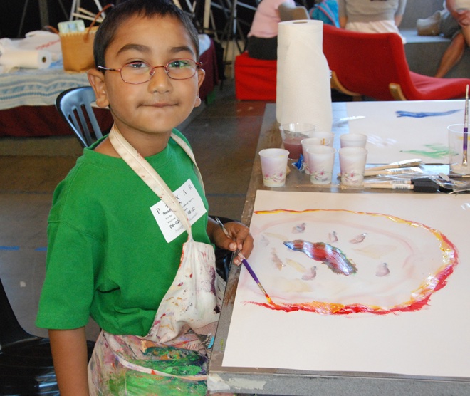 A young boy with short dark hair and glasses is participating in a science art activity. He is wearing glasses, a green t-shirt and an apron splattered with paint. The child is smiling slightly while holding a paintbrush and working on a colorful painting in front of them. The painting features abstract shapes with a prominent central figure surrounded by a circular design. Various art supplies, including cups of water, paintbrushes, and paper towels, are arranged on the table. Other children and adults can be seen in the background, engaged in similar activities.