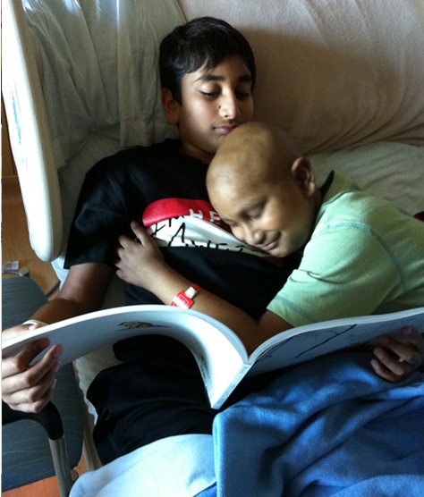 Two brothers are sharing a tender moment in a hospital setting. The older brother, wearing a black t-shirt with red and white graphics, is sitting up in a hospital bed and holding a book open. The younger brother, who has a bald head and is wearing a green shirt, is lying next to him with his head resting on the older brother's chest. The younger brother has a hospital wristband on his wrist. Both boys appear relaxed and content as they enjoy the book together. The background shows typical hospital furnishings, including the bed and some medical equipment.