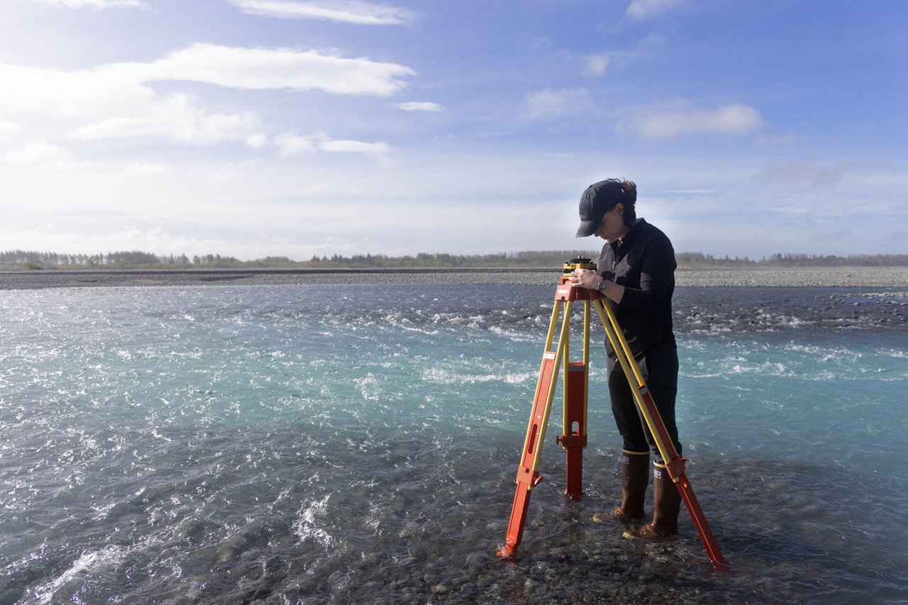 A photo of a field researcher standing on the edge of a river in New Zealand, setting up a GPS unit on a tripod.