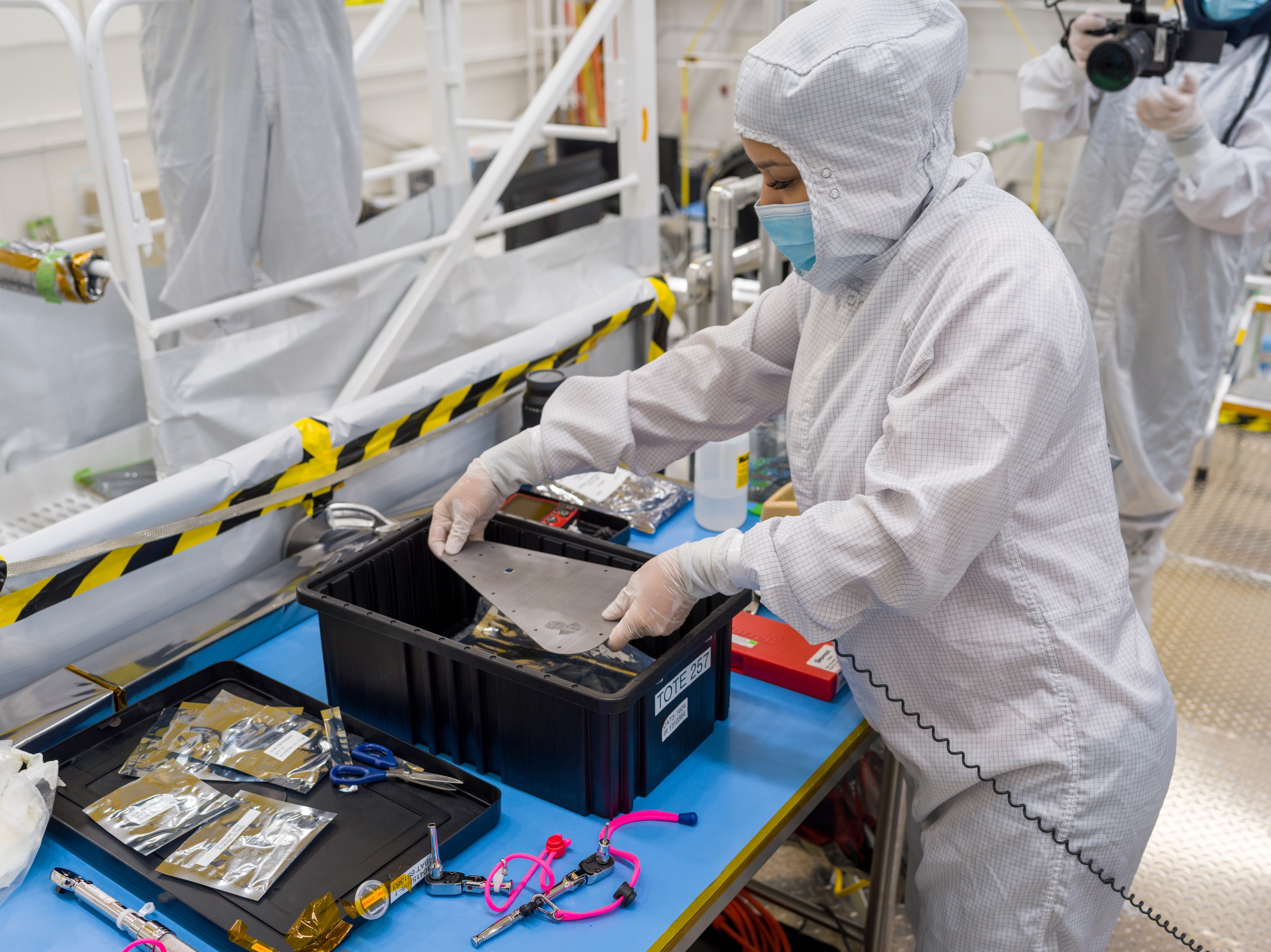A technician in white protective clothing holds the triangular-shaped, silver-colored vault plate over a black box sitting on a work table with lots of tools around it. Another worker in protective clothing holds a video camera to record the moment.