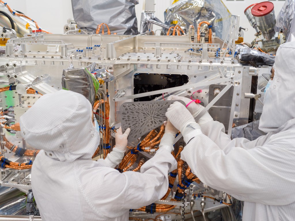 Two workers in white protective clothing hold the triangular-shaped, silver-colored vault plate near the Europa Clipper spacecraft.