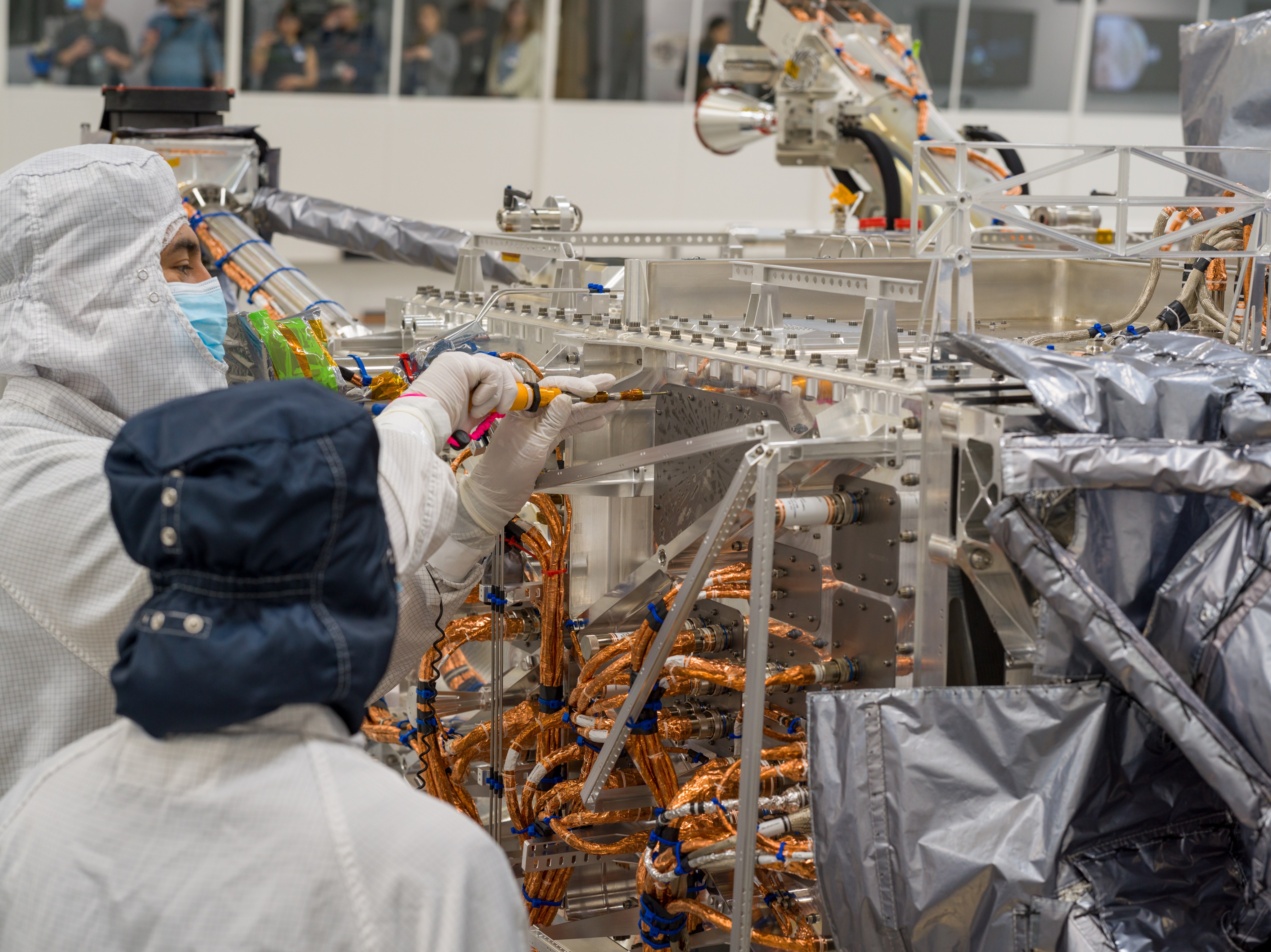 A worker dressed in white protective clothing uses an orange-handled screwdriver to turn a screw attaching the triangular-shaped silver-colored vault plate to Europa Clipper. Wires and other parts of the spacecraft are visible. Another worker dressed in white and blue head cover looks on.