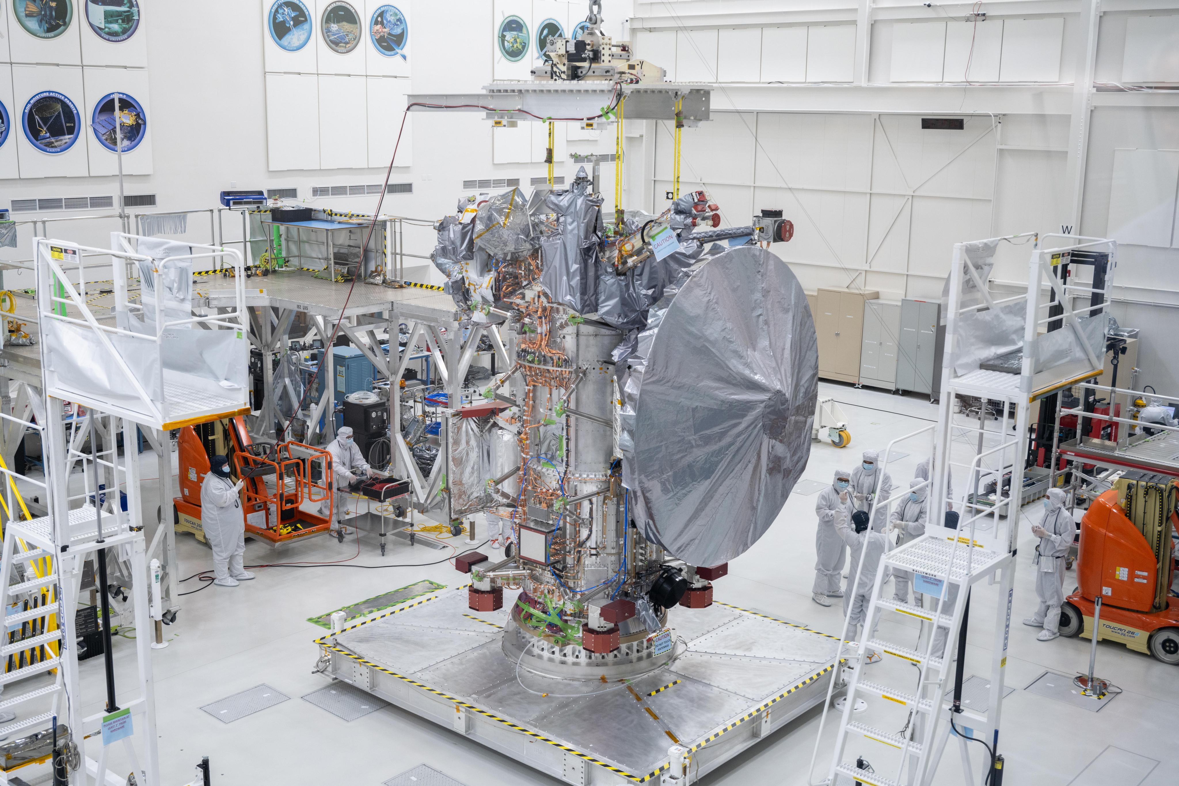 The silver-colored Europa Clipper sits on a work stand in a clean room. Workers in protective clothing stand nearby.