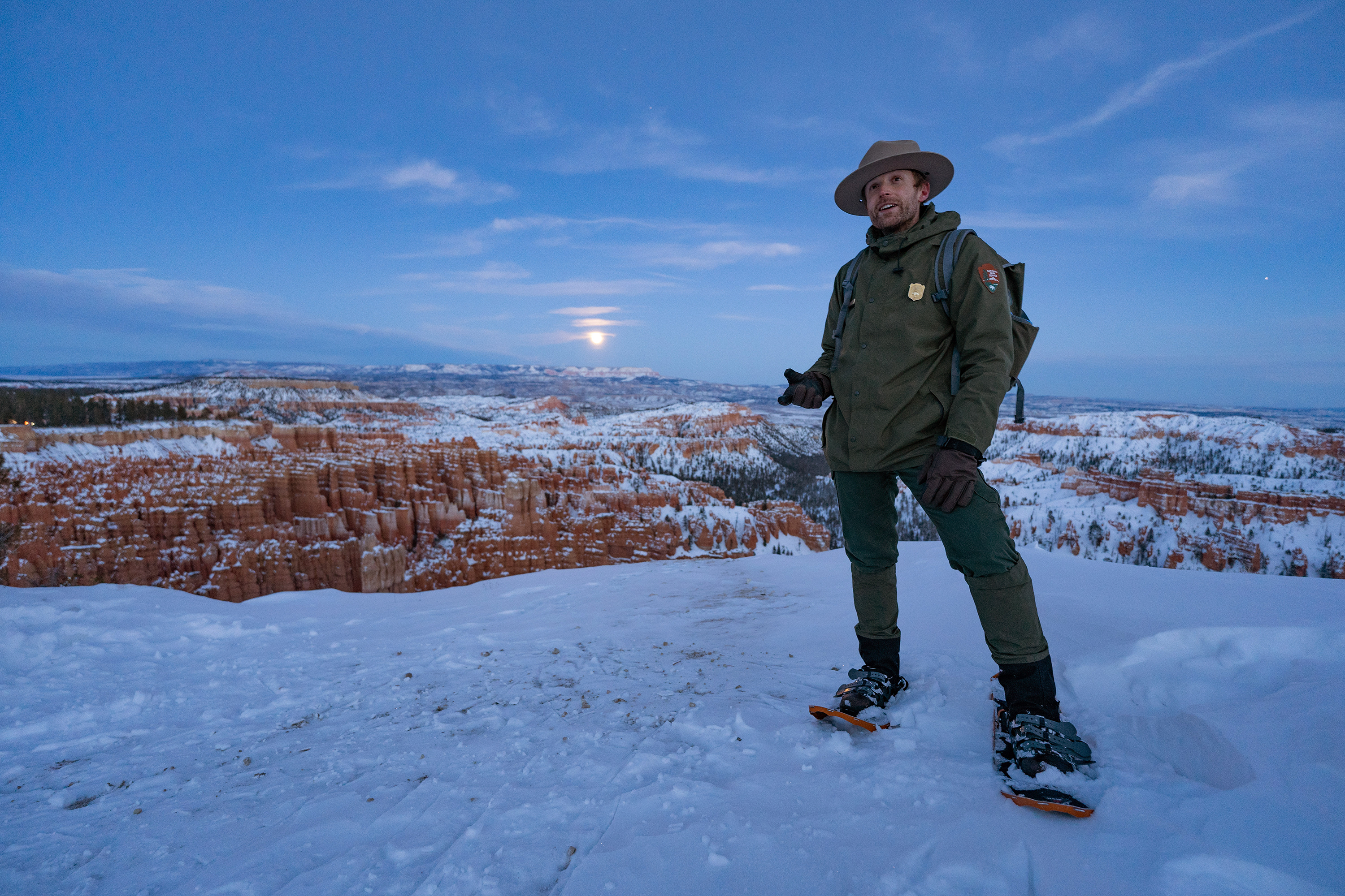 A park ranger stands on a snowy vista pointing to a full moon in the background.