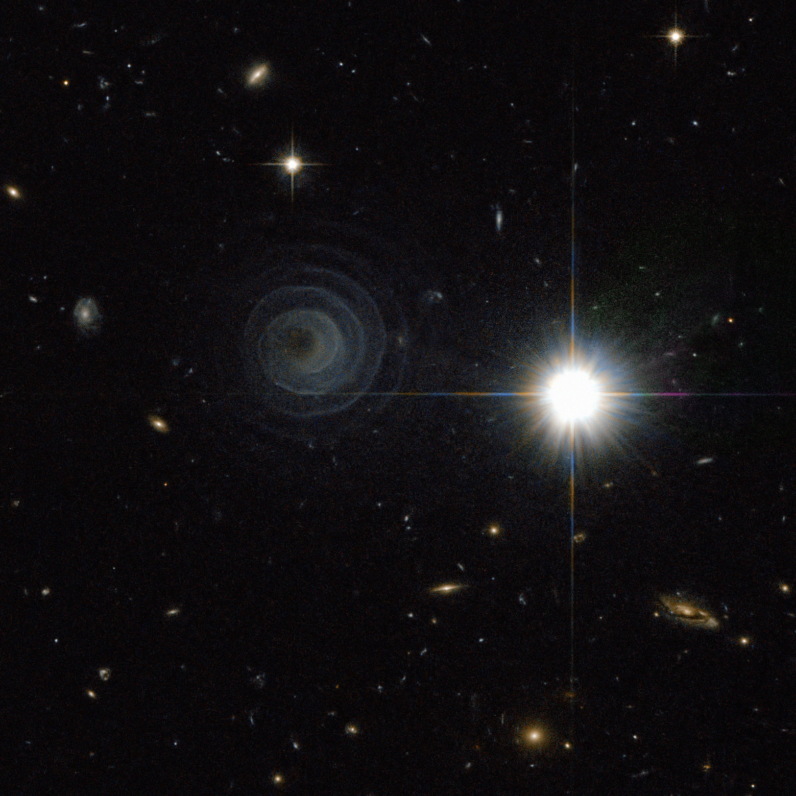 A black background is punctuated with distant stars and galaxies. One bright foreground star sits just to the right of image center. Slightly above and to the left of image center is a faint, bluish-green spiral.