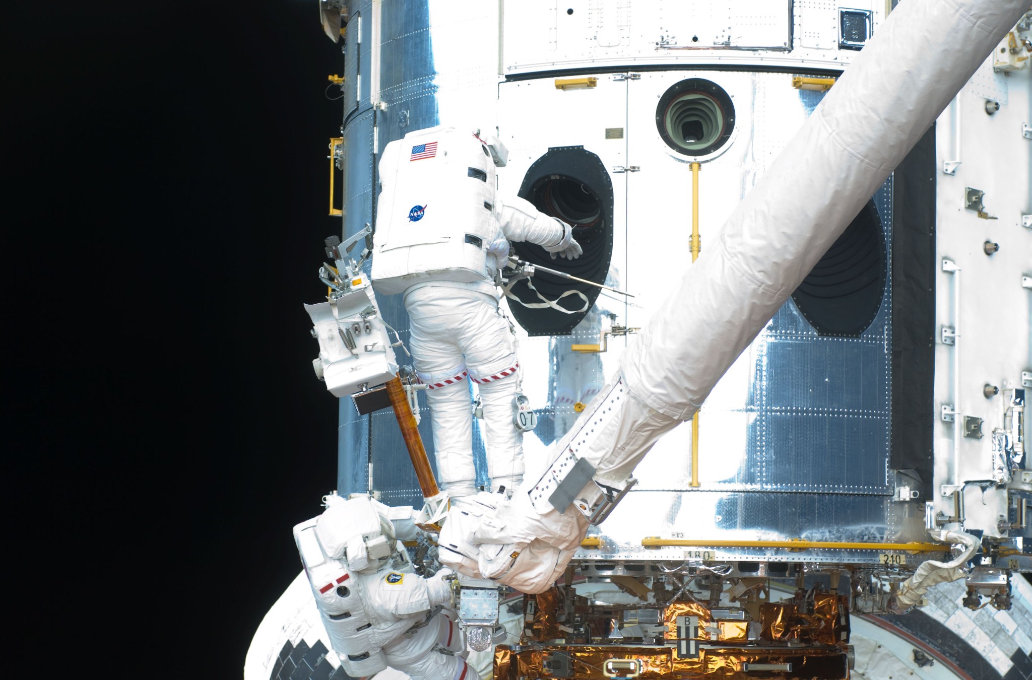 Two astronauts, Mike Massimino assists Michael Good who is standing on the Manipulator Foot Restraint, which is connected to the Shuttle Remote Manipulator System/Canadarm