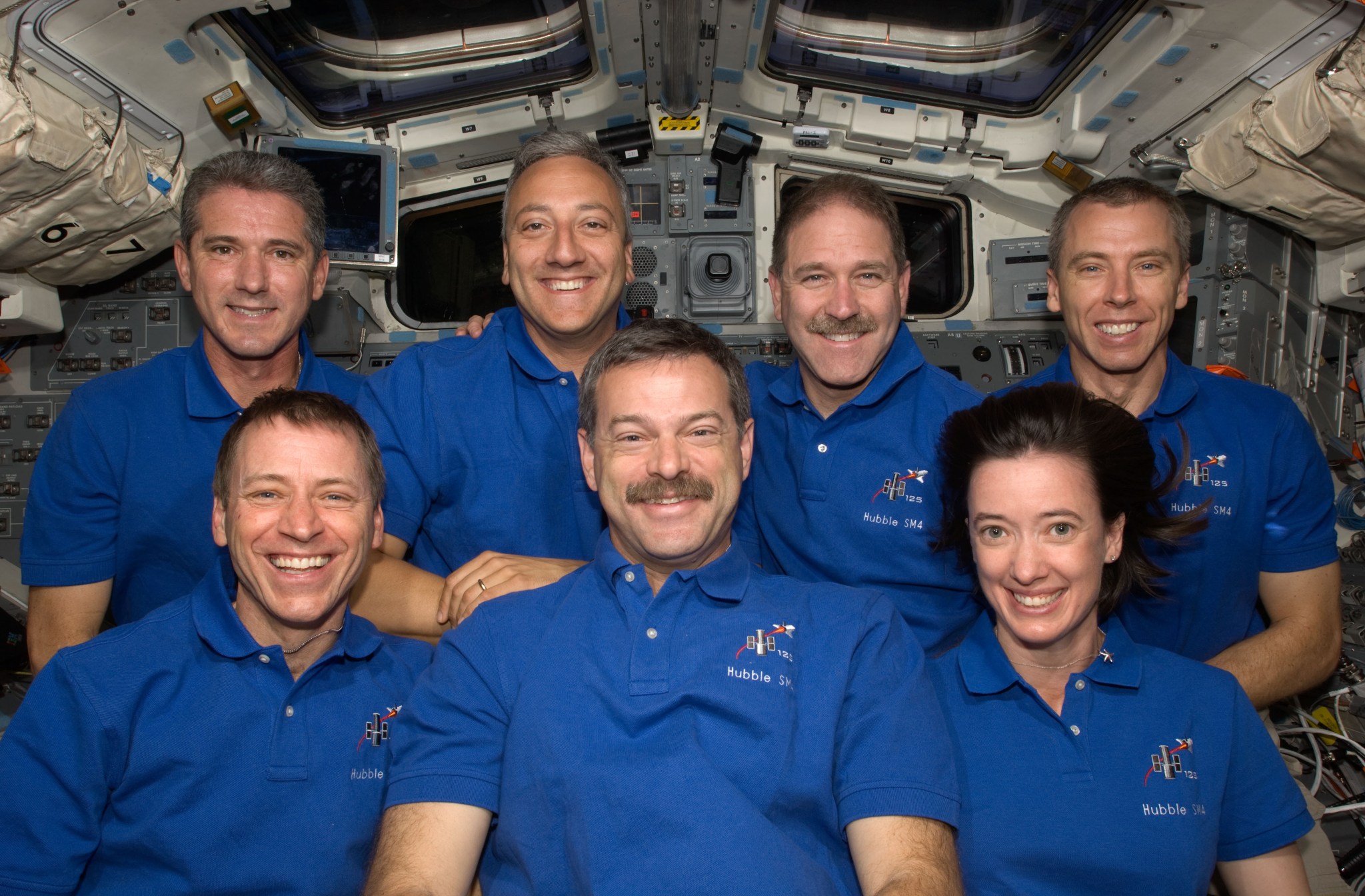 STS-125 crew poses in front of an American flag in the crew cabin of the shuttle.