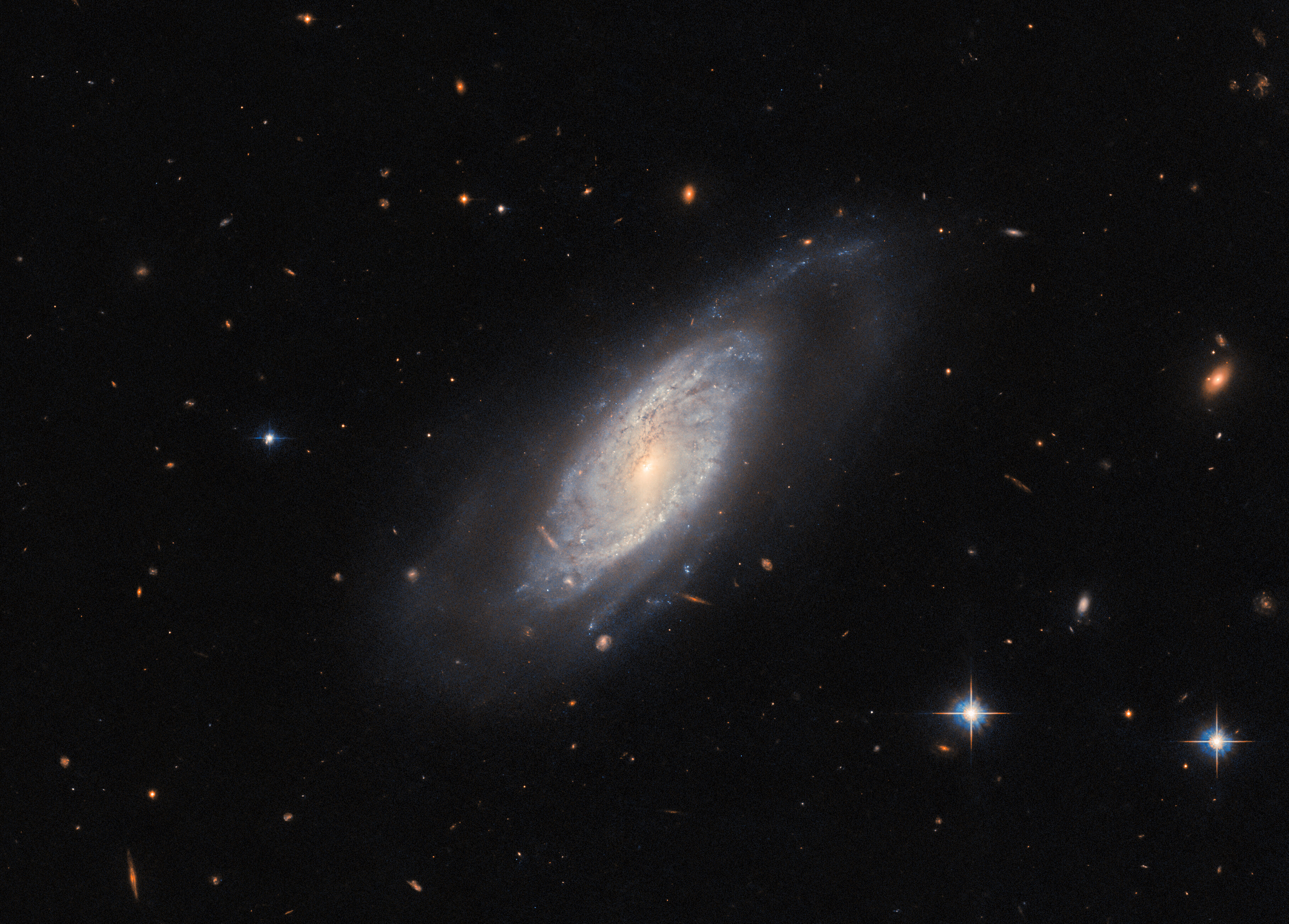 A spiral galaxy in the center of a dark background, surrounded by a few distant galaxies and nearby stars. The galaxy stretches diagonally across the center of the frame — from lower left to upper right — and is slightly tilted toward the viewer. Its cloudy disk is threaded with dust and holds no clear arms. A bar of light extends across the disk from the glowing core. A faint halo of gas surrounds the disk.