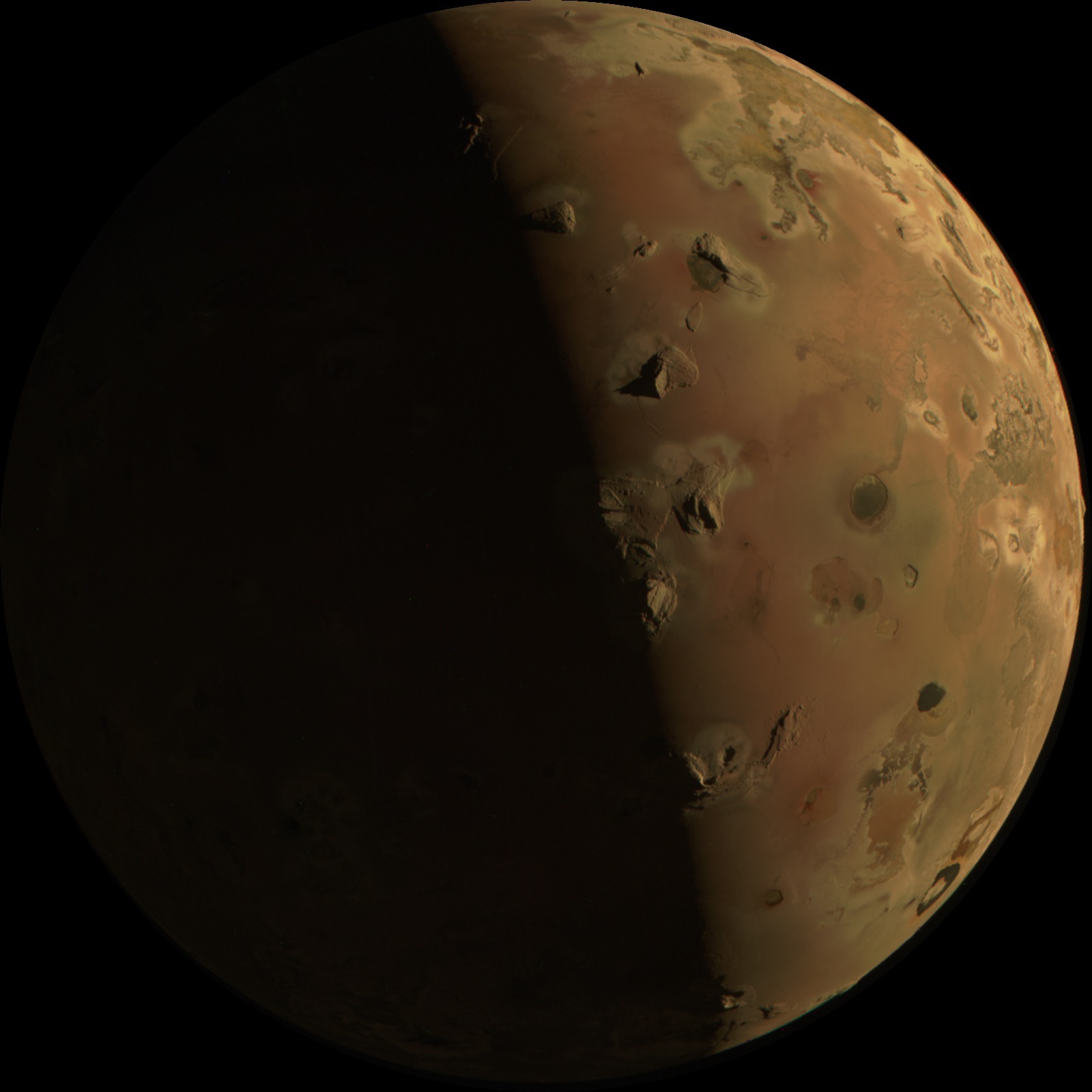 A rust-colored sphere is shown against a black background. The left half is concealed in shadow, with only a very dim outline visible. The right half is fairly well-lighted, with the surface smooth in some areas and in others covered with splotches and peaks of light tan, or spots and dimples of dark orange or dark grayish brown.