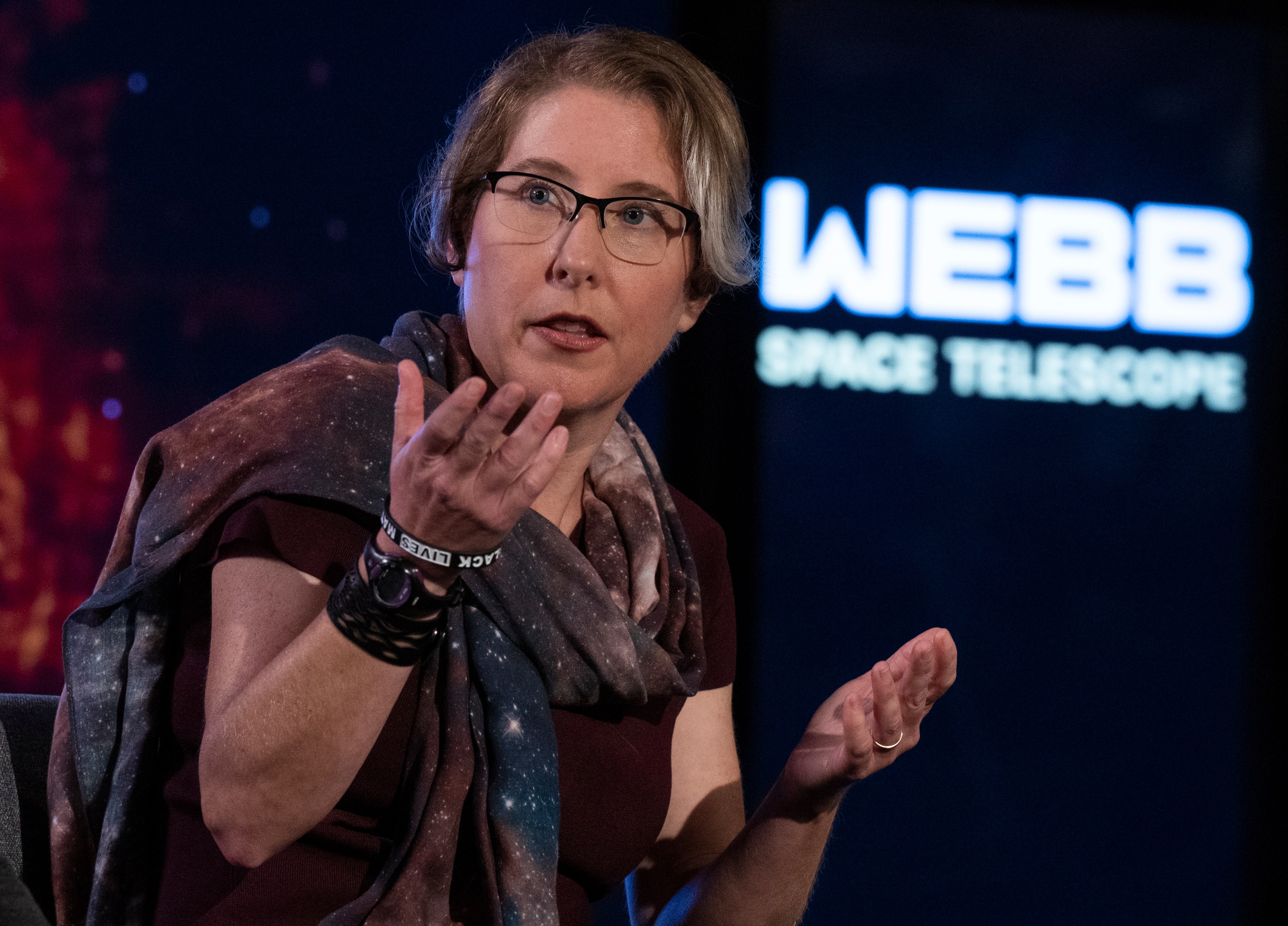 A fair-skinned woman with short light brown hair, wearing a chunky galaxy scarf, and thin-framed glasses looks off to the side, speaking with her hands. Behind her is faded text that says &quot;WEBB SPACE TELESCOPE&quot;