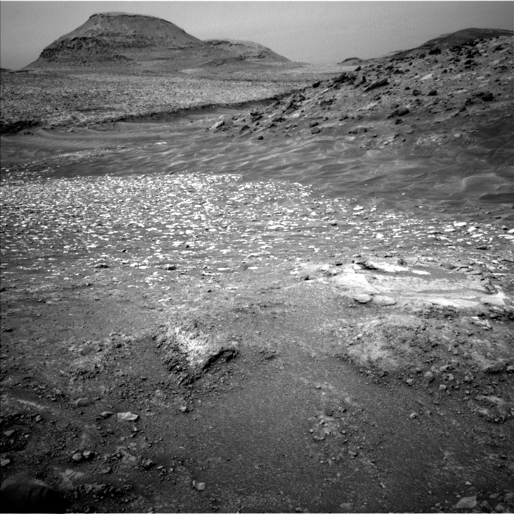 Black and white image of Mars taken by Curiosity. There are san dunes leading up to a few rocky hills in the background.