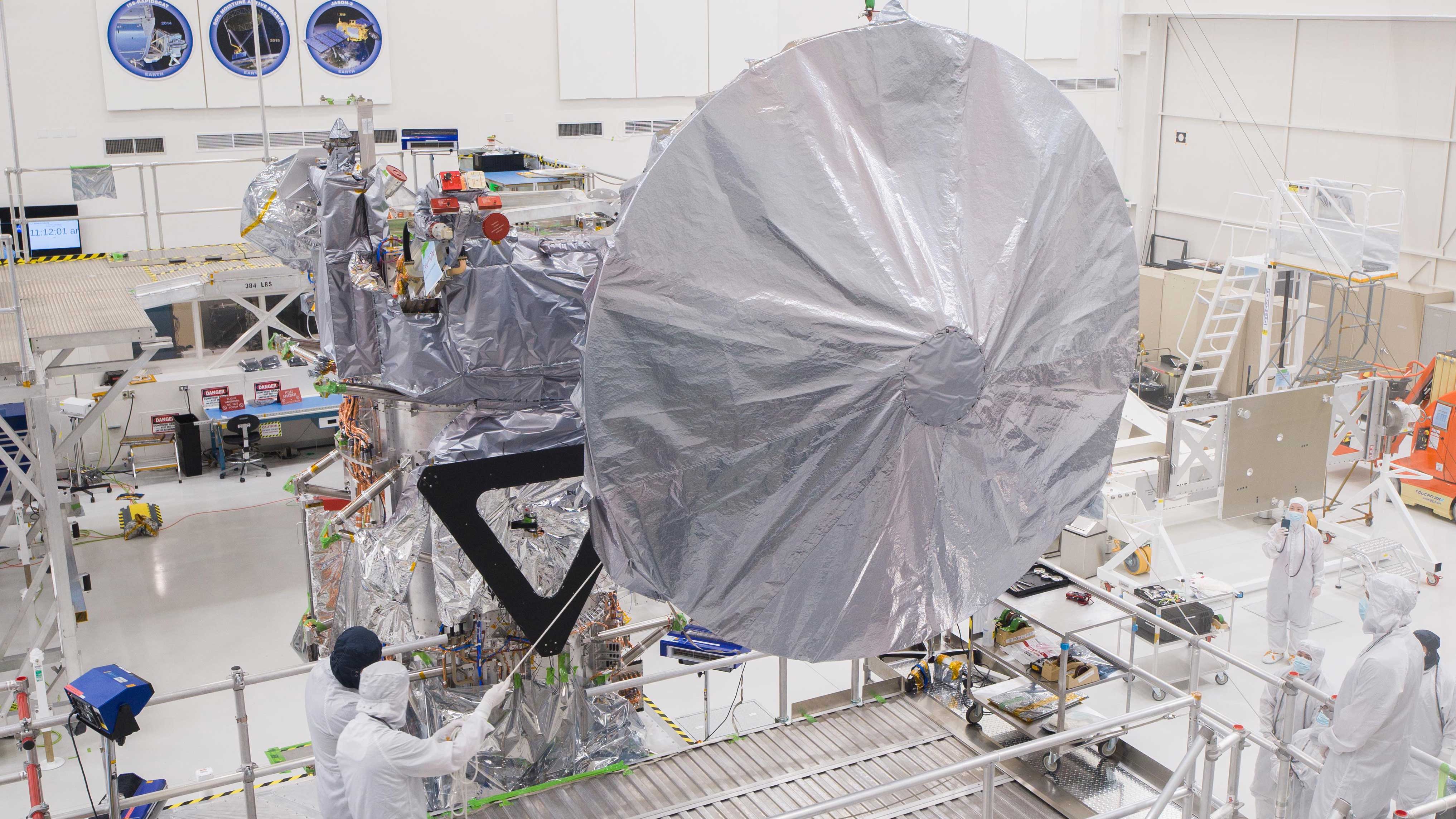 Workers in white protective clothing lift the silver, disc shaped Europa Clipper high gain antenna.