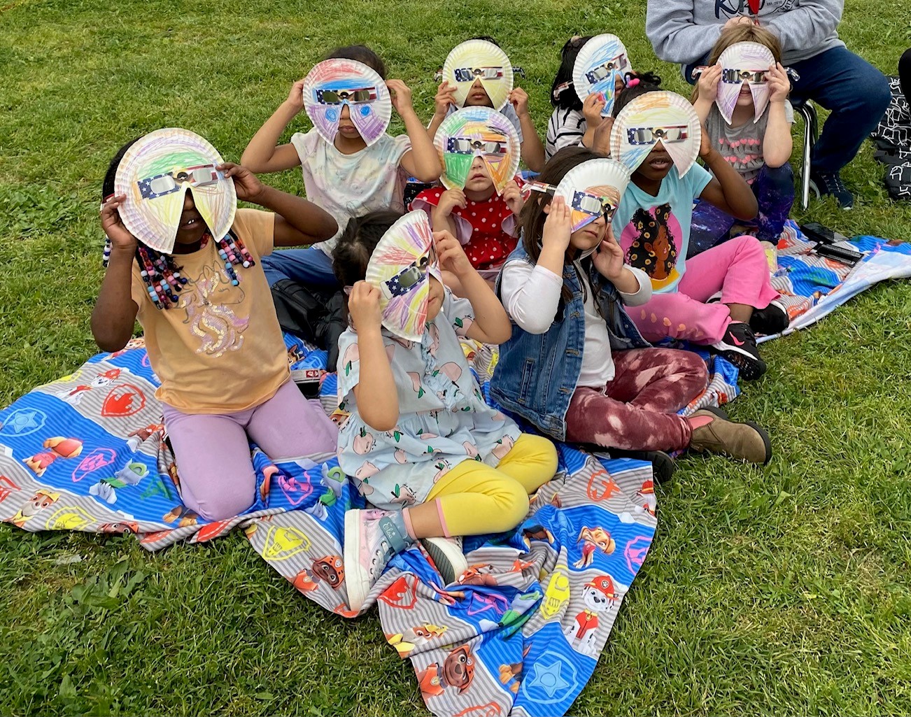 Nine young children sitting on a blanket outside using paper plate eclipse viewers to see the sun.
