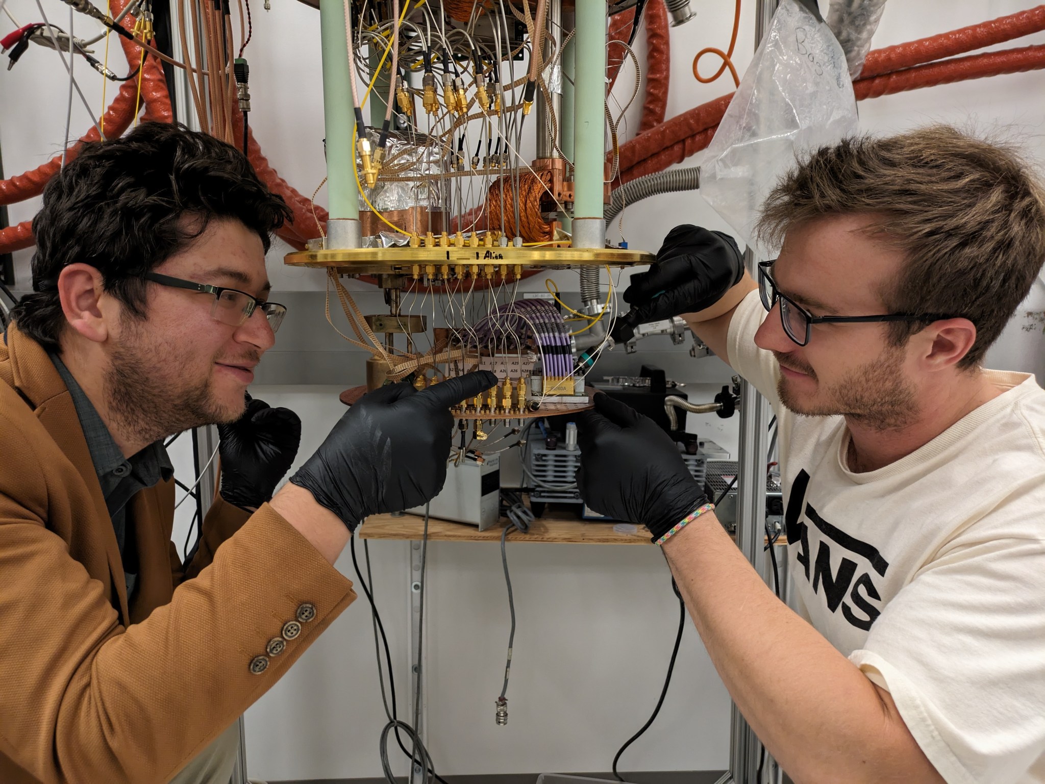 Two NIST team members stand beside an exposed cryogenic refrigerator, wearing gloves while affixing an aluminum sample box with wires coming out of it to a copper sample stage