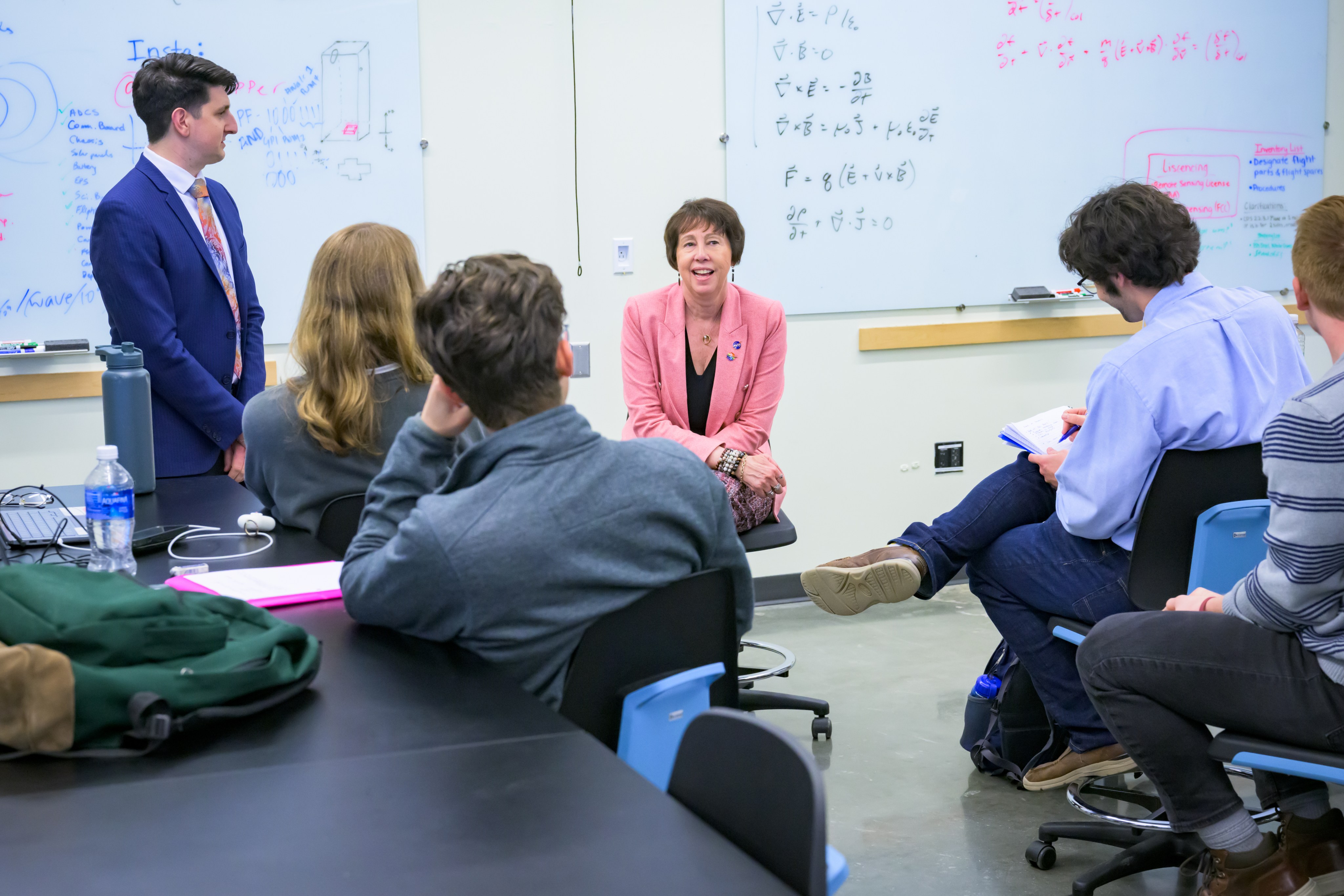 Dr. Nicky fox casually chats with undergraduate students over coffee in a laboratory at the University of Delaware. Students take notes in front of a whiteboard while seated around Dr. Fox.