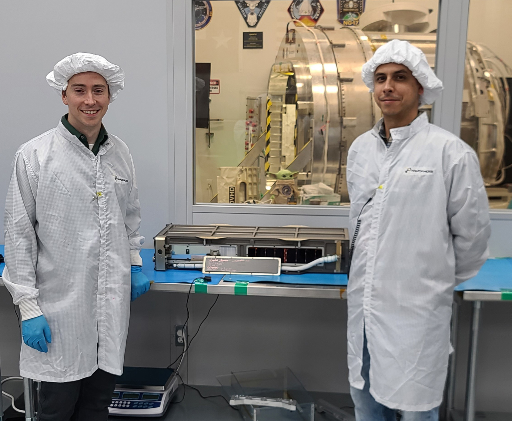 Two people wearing white lab coats standing beside a small, box-shaped satellite in a laboratory