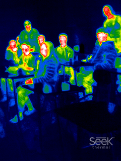 This is a thermal image depicting several people gathered, likely indoors. The varying colors represent different temperatures, with warmer areas shown in red and yellow and cooler areas in green and blue. The people's heads and other body parts that are emitting more heat appear in warmer colors, indicating they are warmer than the surrounding environment.