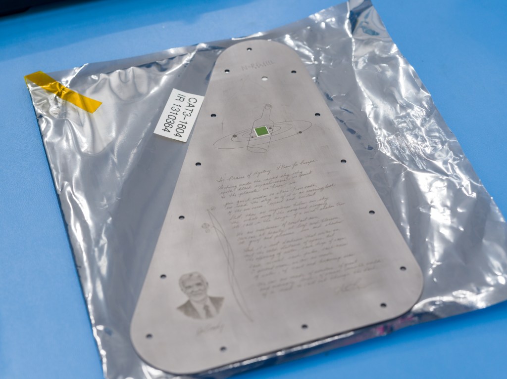 The triangular-shaped, silver-colored Europa Clipper Europa Clipper vault plate is covered in a protective plastic bag with a clear front and silver back. There is a bit of gold tape in the upper left corner, and a white label on the bag.