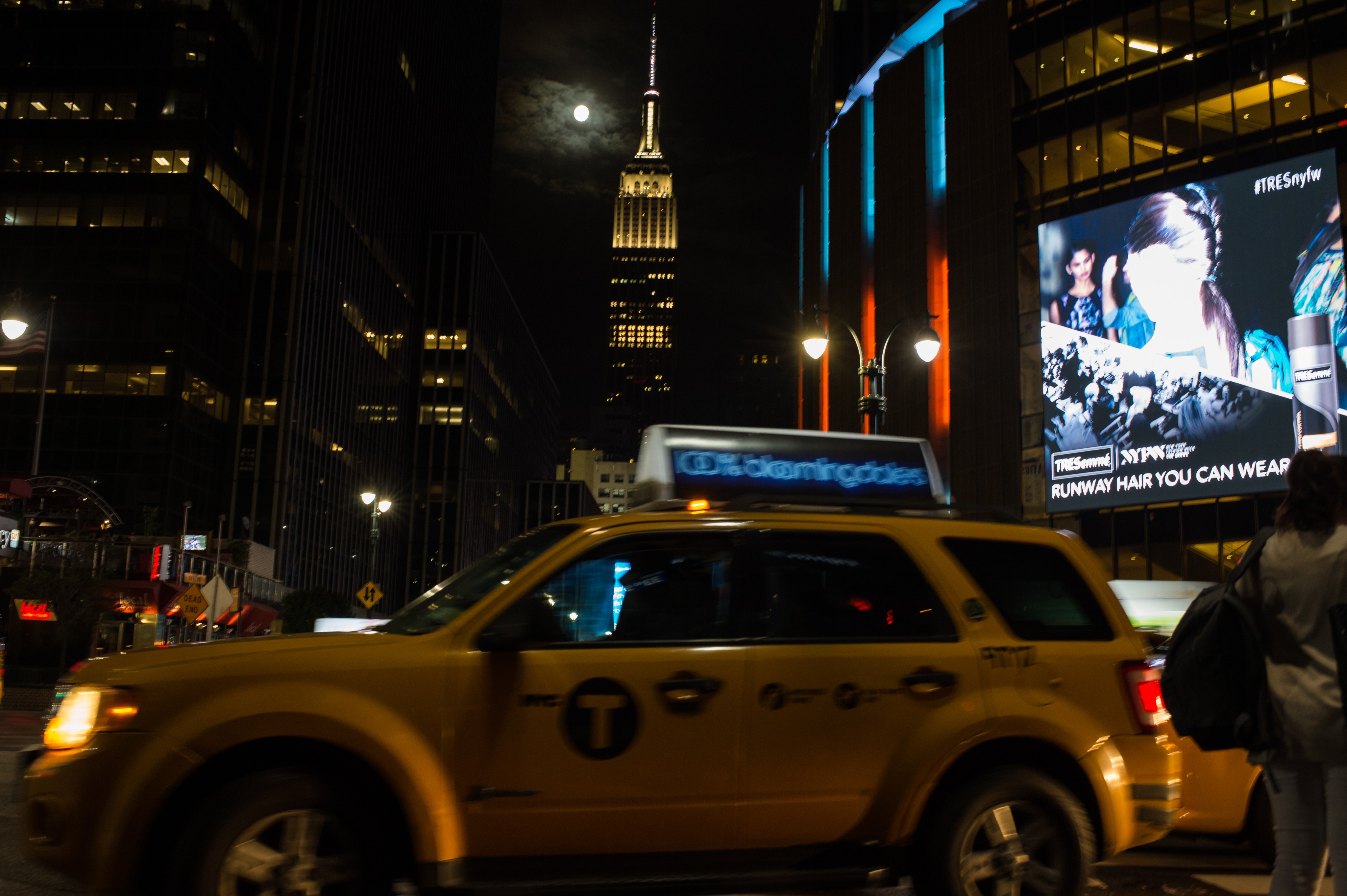 The full moon is visible next to the Empire State Building in this photo of New York City. A cab is rushing across the image in the foreground.