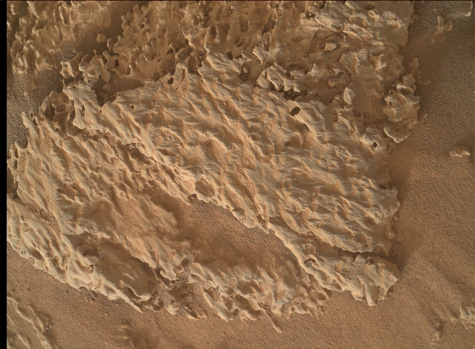 Pale orange terrain on the surface of Mars, with a sandy area creating a reverse "L" shape anchored in the lower right corner of the frame and extending to the upper right and lower left corners. The rest of the frame is covered in uneven, rocky ground of the same color that resembles meringue or whipped, peaked frosting spread on a cake.