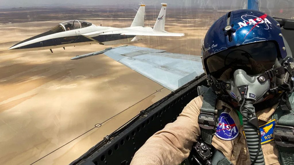 A pilot sits in the cockpit of a plane wearing a blue helmet with the NASA logo, and a pilot oxygen mask. Another plane can be seen off to the pilot's left side.