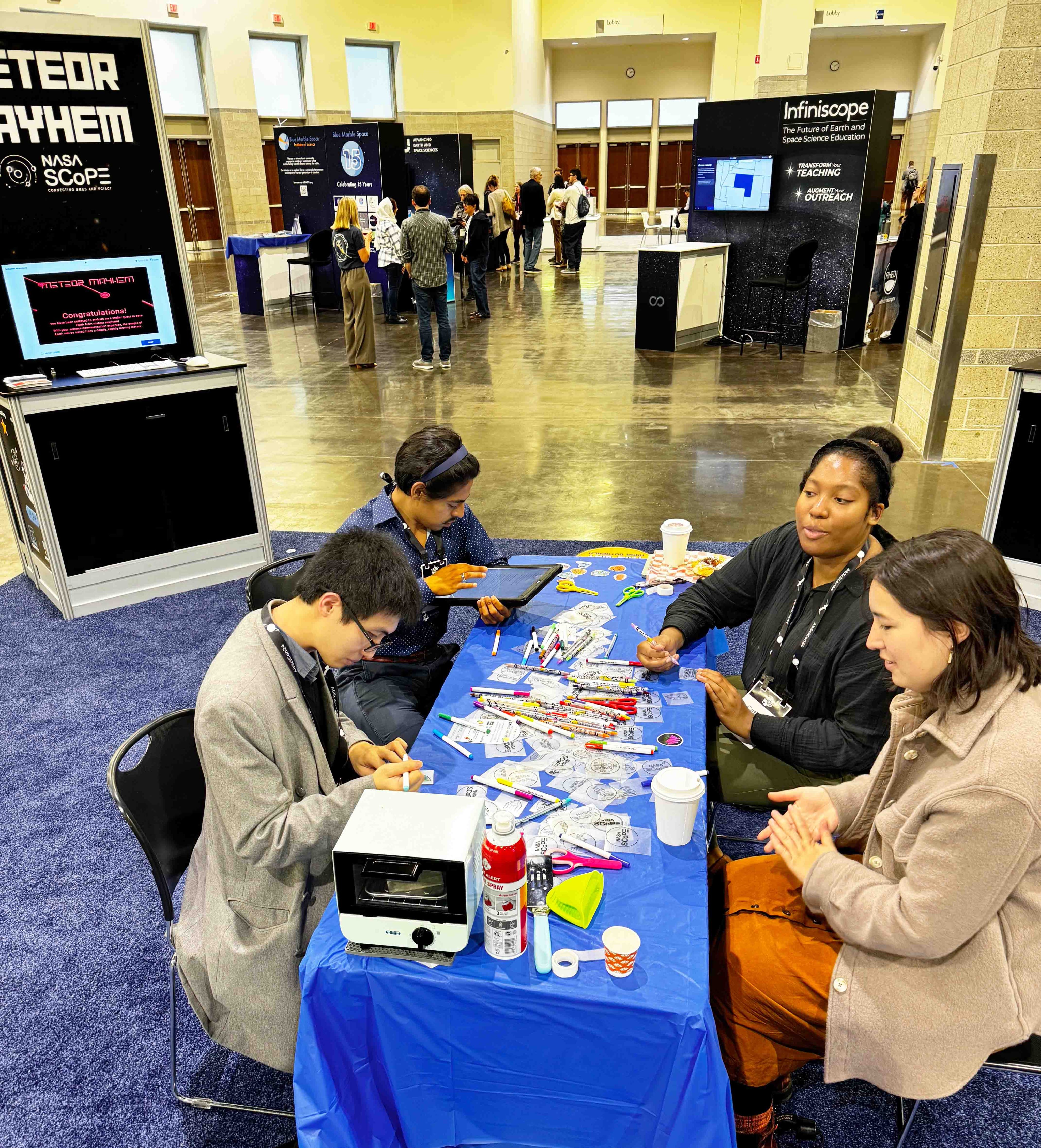 A group of four people, sitting around the exhibit hall table, engaging in a shrinky dink craft. The table is covered with a blue cloth and various materials including pens, badges, and small tools. Behind them are exhibit booths labeled "Meteor Mayhem" and "Infiniscope," with additional conference attendees visible in the background.