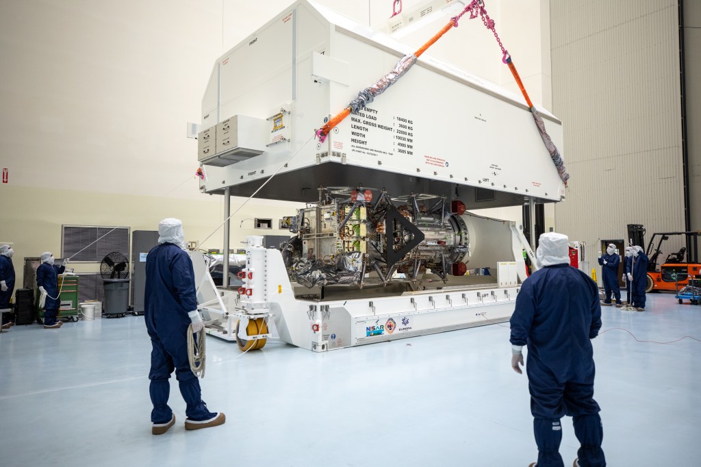 Workers in blue protective clothing and white head covers watch as Europa Clipper is lifted and rotated.