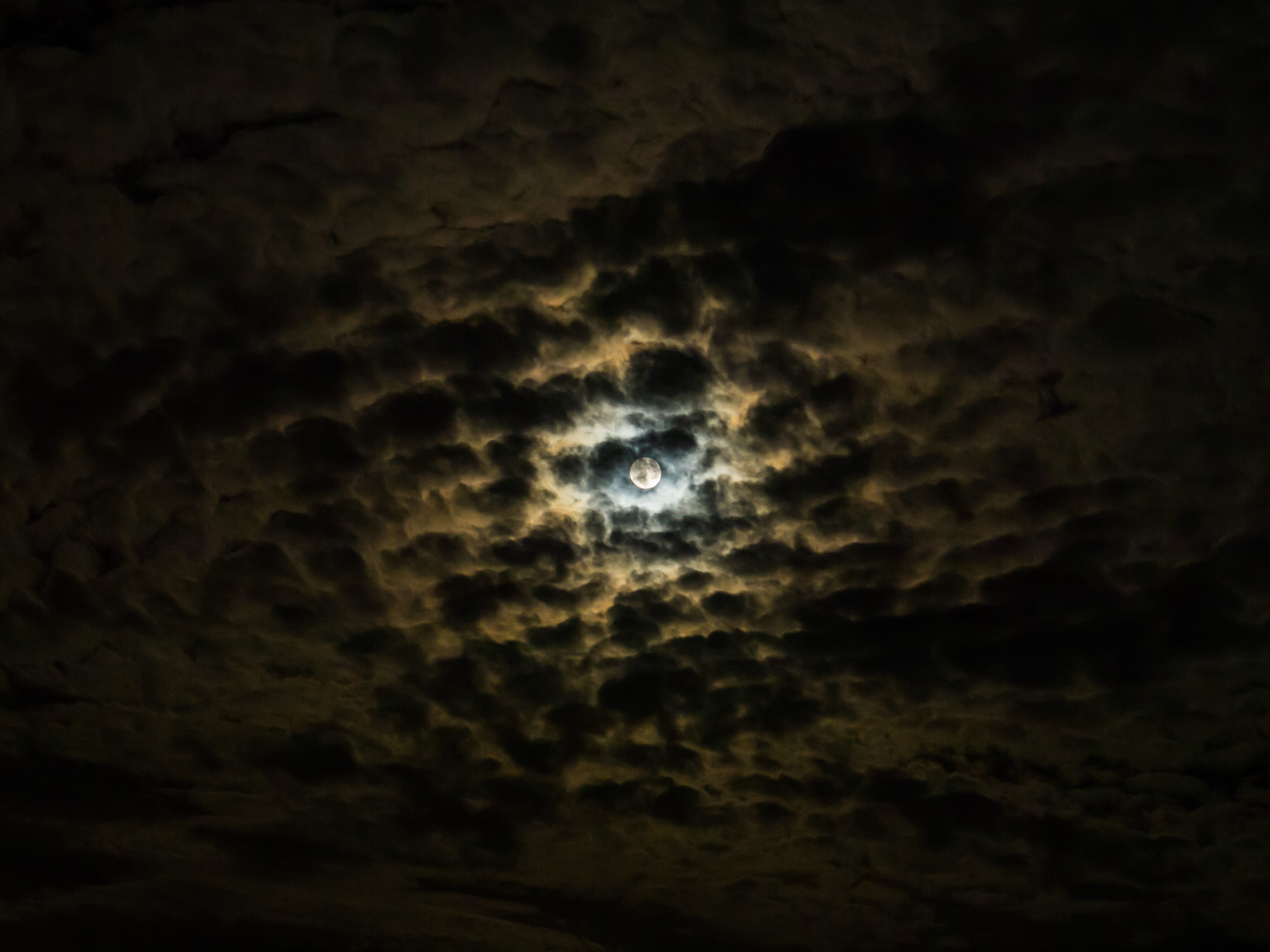 The Moon is seen behind puffy black and orange clouds at night.