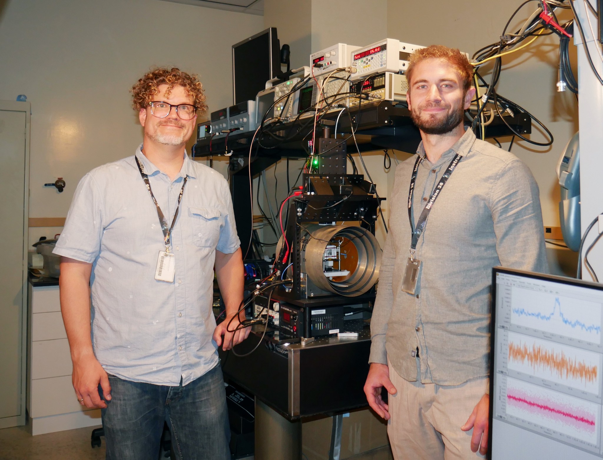 Two smiling team members stand on either side of an optical table with multiple instruments and a barrel-shaped open chamber.