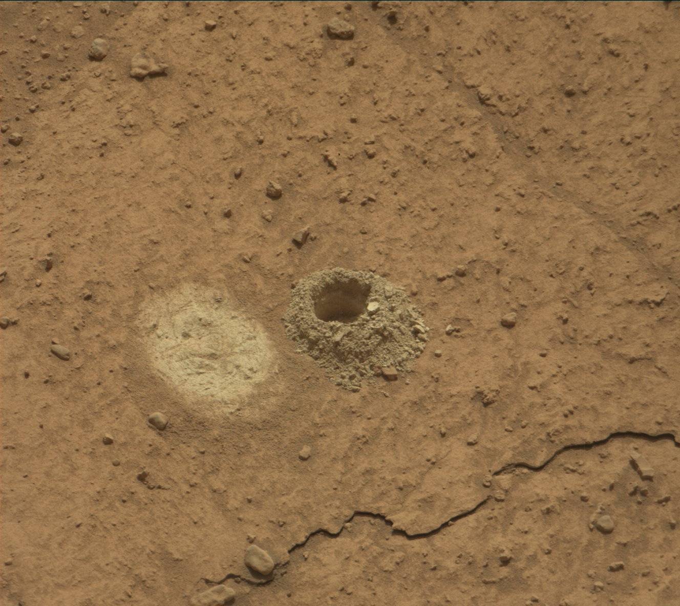 A patch of dry, hard, barren, orange-tan ground fills the frame, with a pronounced crack in the ground running from the lower-right to the bottom-center of the frame. In the middle is an oval patch in light tan, where the top soil has been scraped away, and to its right is a small hole in the ground looking like a tiny crater, with soil dug out of the hole crating a sloped wall around its edges.