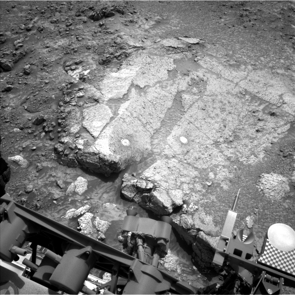 A black and white photograph of the Mars surface, with some of the metallic structure of the Curiosity rover visible at bottom of the frame. Most of the upper three-quarters of the frame shows an area of flat, broken slabs of light-gray rock in front of the rover, surrounded by darker areas of soil and gravel. The slabs have two small, whitish, ellipitacal marks on their surfaces.