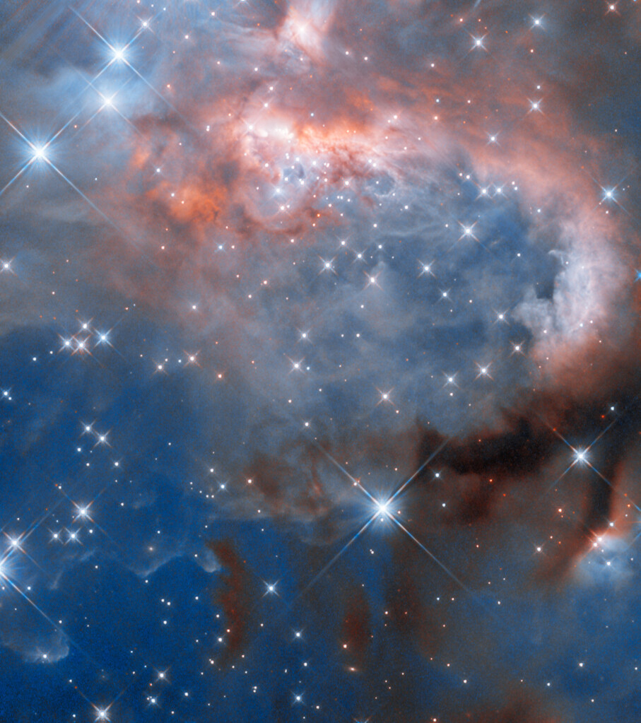 Clouds of gas and dust with many stars. The clouds form a flat, blue background toward the bottom, and become thicker and smokier toward the top. Stars on one side light the nebula. A thick arc of gas and dust reaches around from the bottom-right corner of the image toward the top-left corner. It begins as a dark and obscuring cloud at bottom right and gradually becomes brightly lit by many stars at the upper left. Other large, foreground stars lie between the nebula and the viewer.