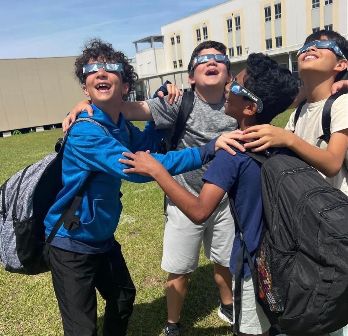 Four young latinx students wearing solar viewing glasses and and looking up at the sun, grasping each other in a circle as if they're jumping up and down with excitement.