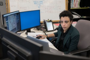 A person with their right hand on a computer mouse, is sitting at a table with five screens in a semicircle around them. The person, wearing a headset, is turned toward one of the screens with a serious, focused expression on their face. They are sitting in a bright office area, wearing a dark dress shirt and blazer.